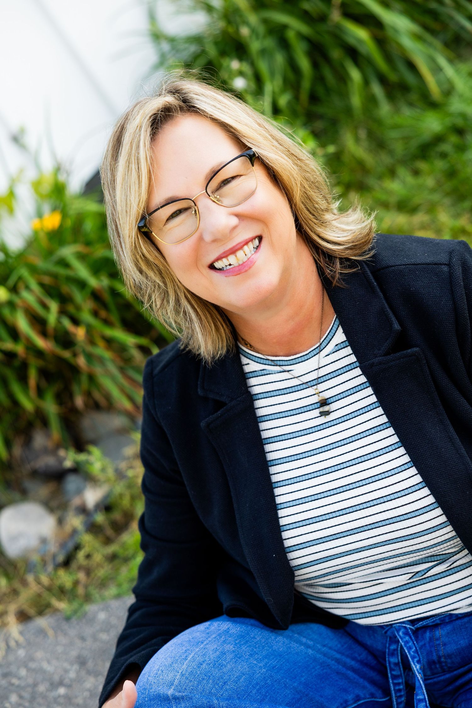 Smiling middle-aged blonde woman with glasses wearing a striped shirt and black blazer sitting outdoors.