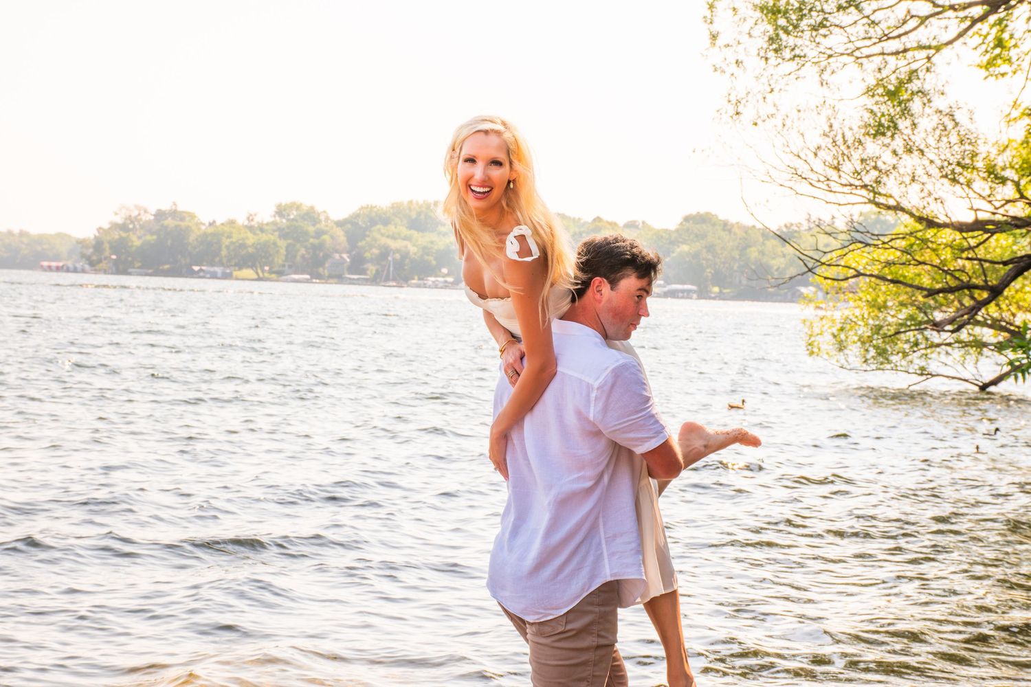 A couple laughs near a lake, man carrying woman on his back in golden sunlight near a tree.