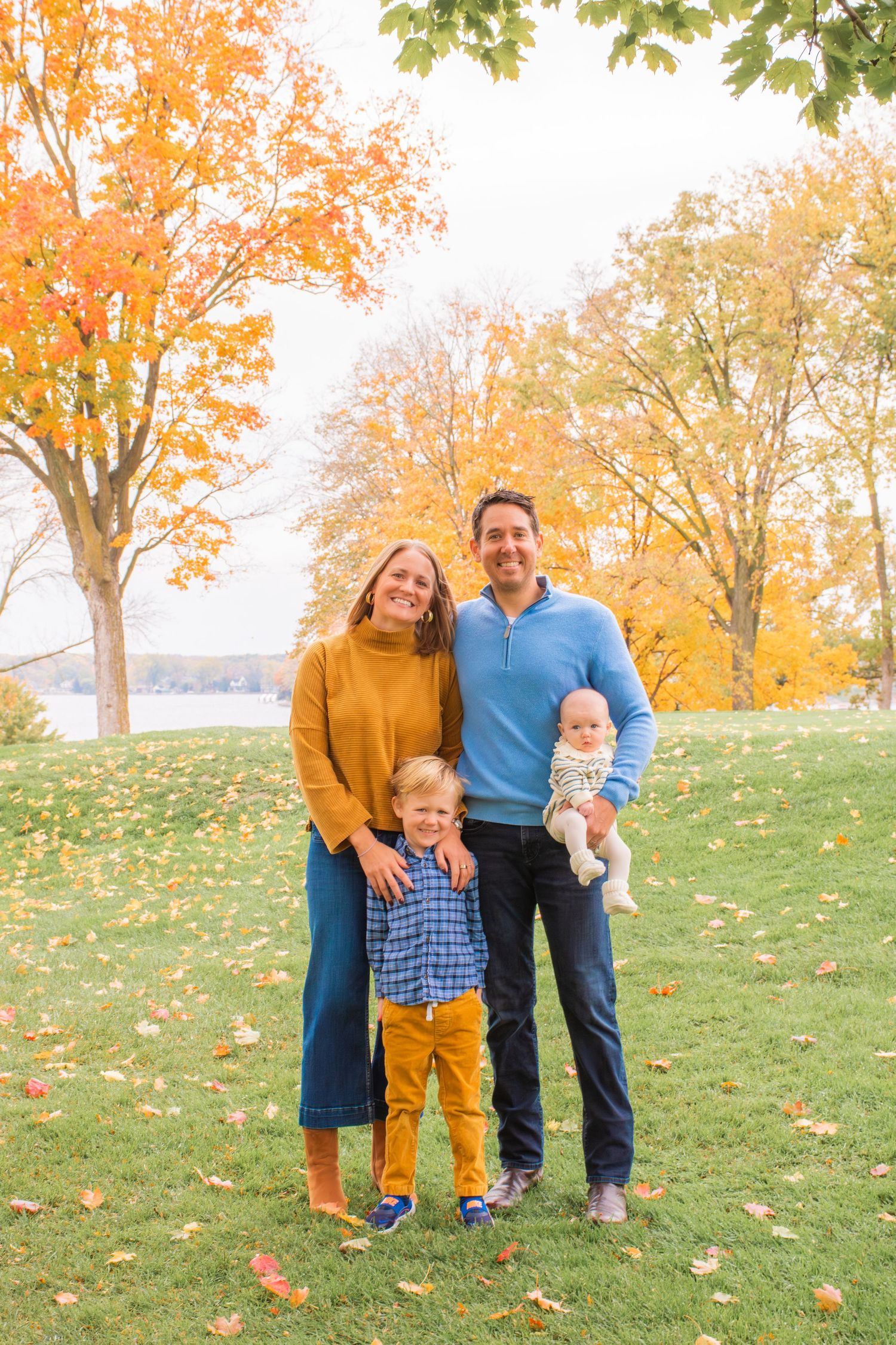 Family of four posing outdoors in autumn park with colorful fall foliage, parents holding baby and toddler son.