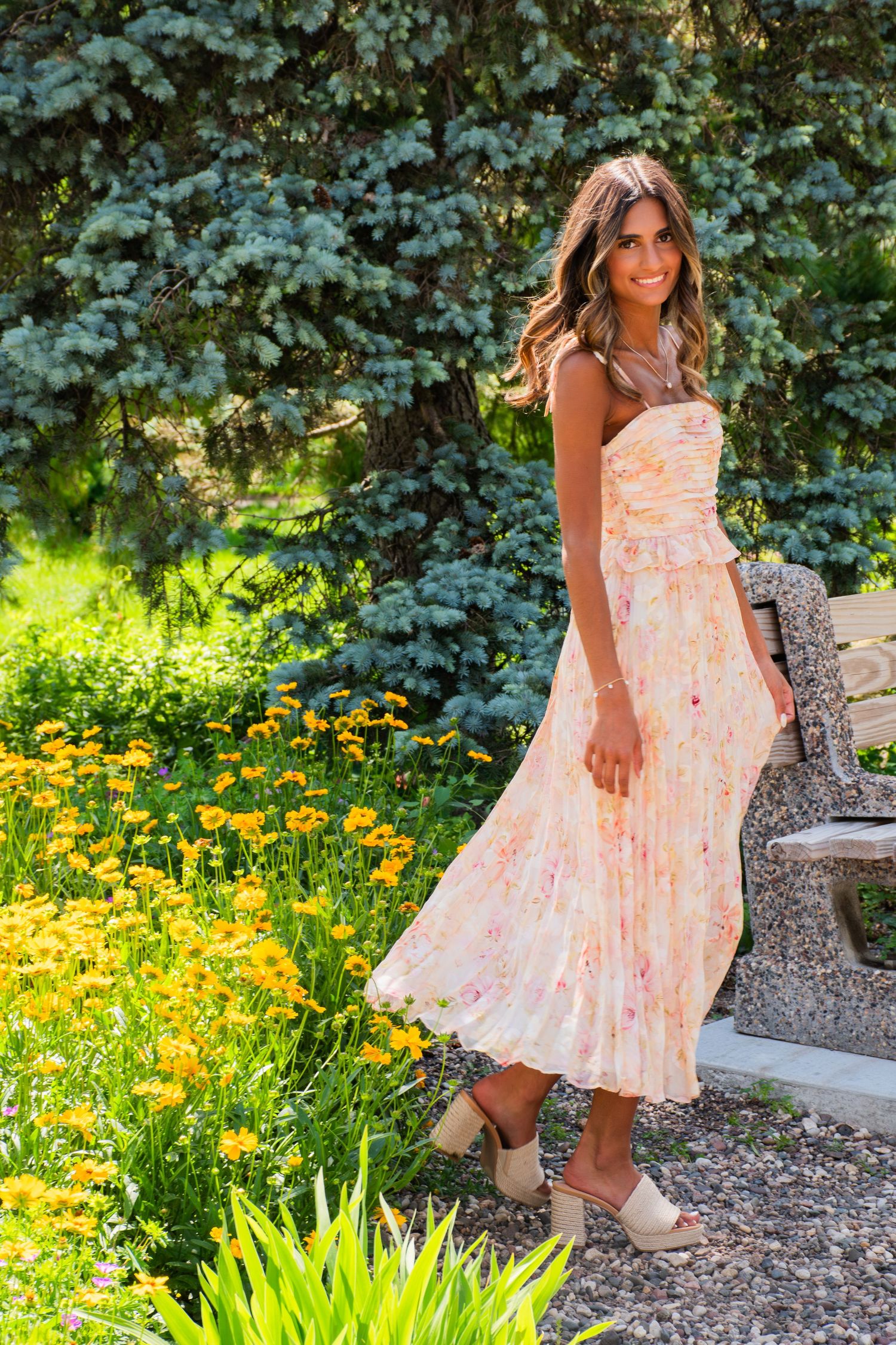 Woman in floral midi dress standing near yellow flowers and stone bench in sunny garden.