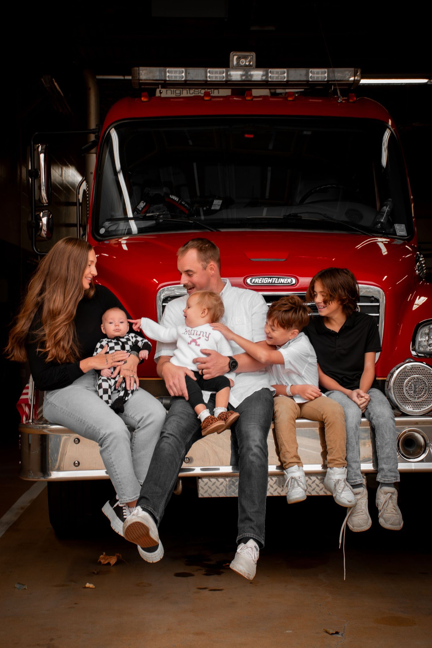 Family of five sitting on front bumper of red fire truck in garage, parents holding babies with older children beside them.