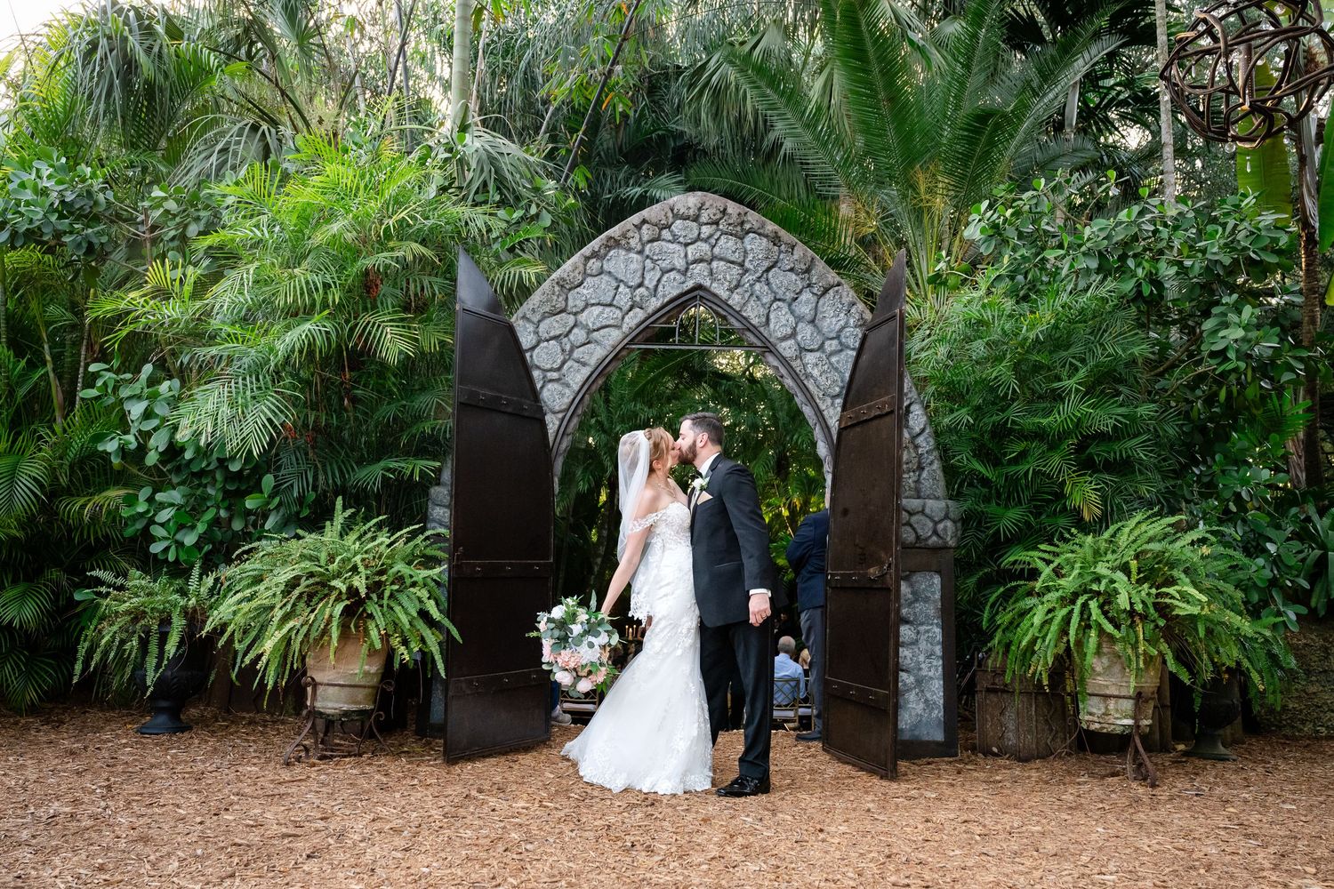 Bride and groom posing beneath a stone arch surrounded by tropical greenery
