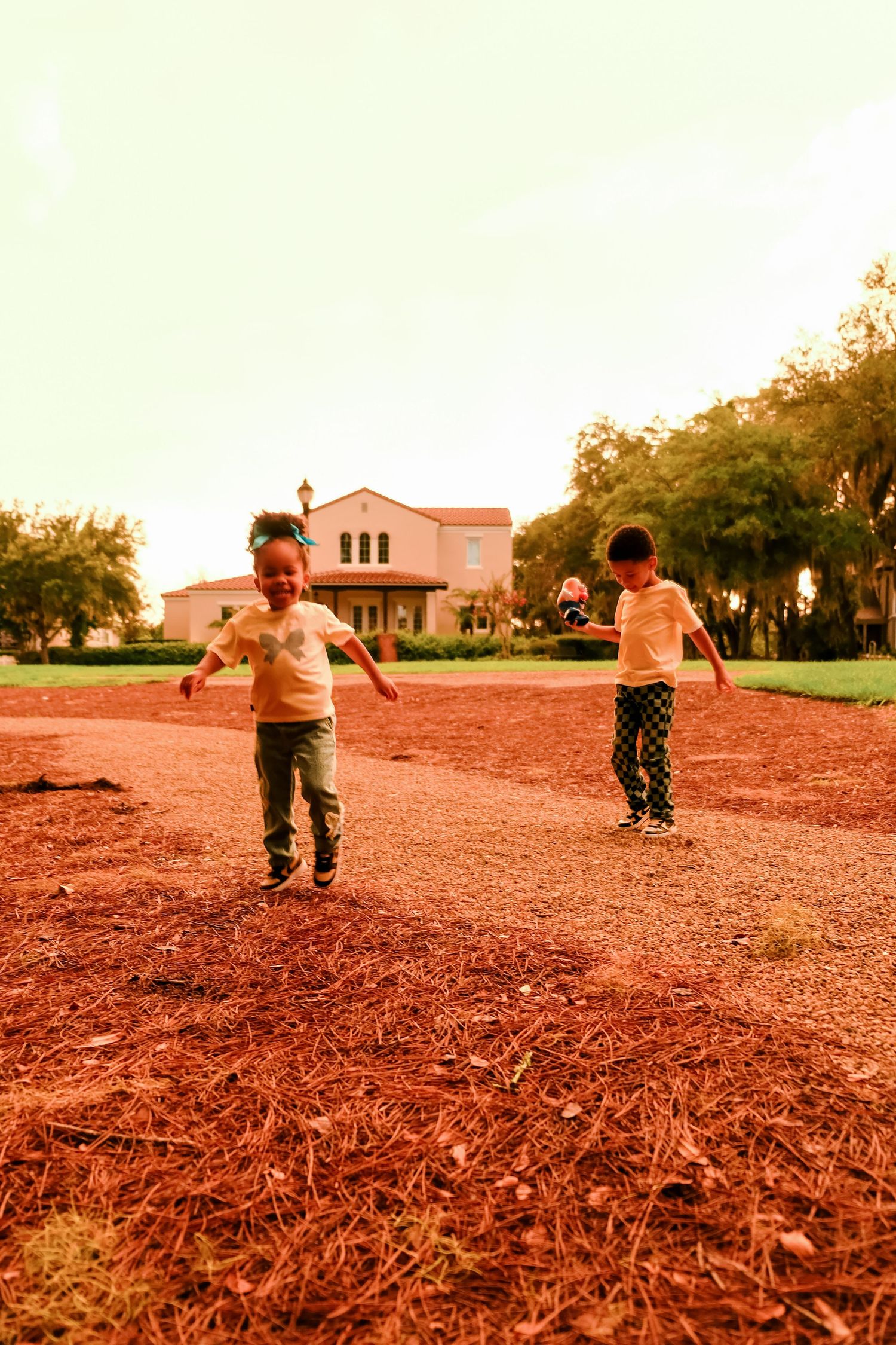 Two children running and playing outdoors on red mulch near a house at sunset.