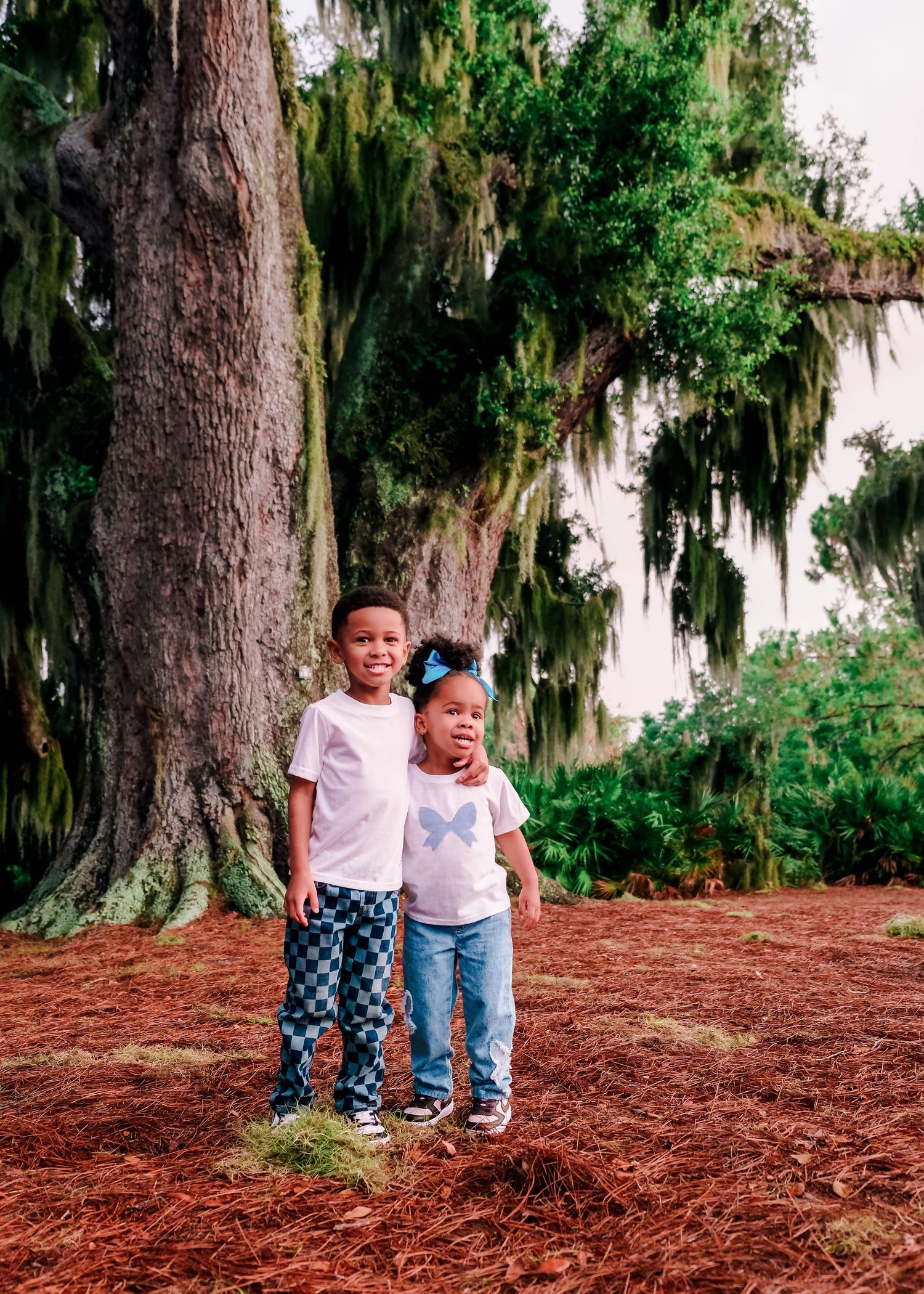 Two children pose in front of a large moss-covered tree on a red mulch ground in a lush green park.