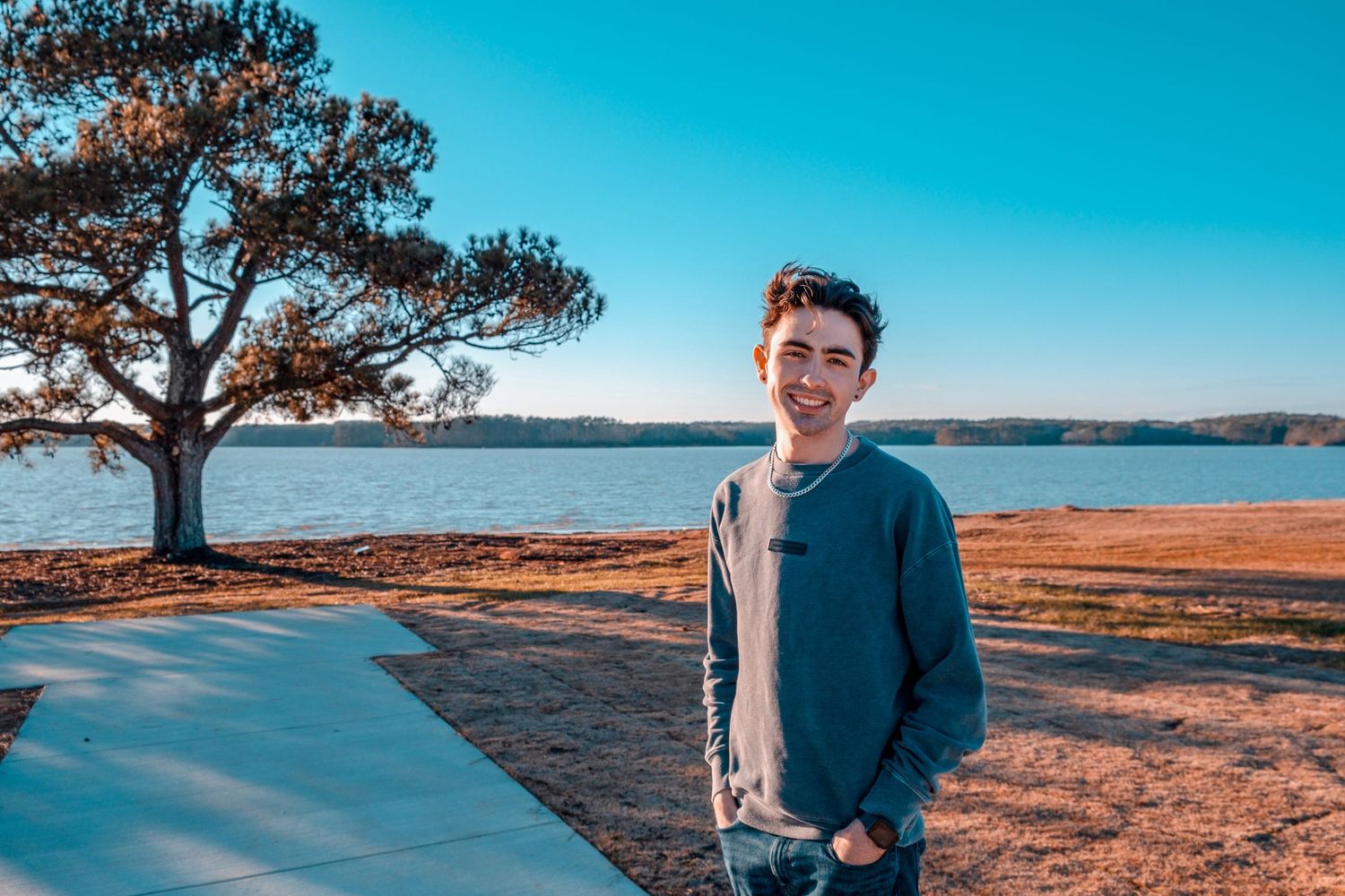 Smiling teenage boy standing near a lakeside path with a tree and blue sky behind him.