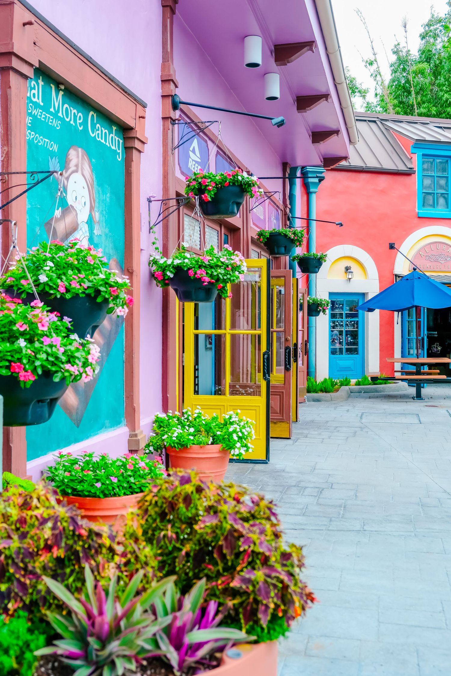 Colorful tropical shopping street with vibrant pink and teal storefronts adorned with hanging flower baskets.