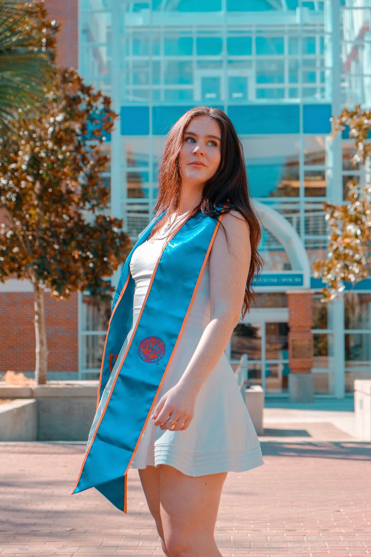 Young woman in white dress and teal graduation sash posing outdoors near a modern university building on a sunny day.