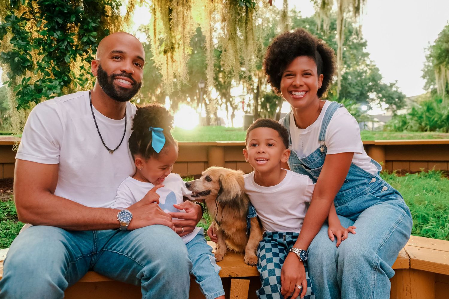 Happy family of four with a dog sitting on a bench outdoors surrounded by lush green trees in warm sunlight.
