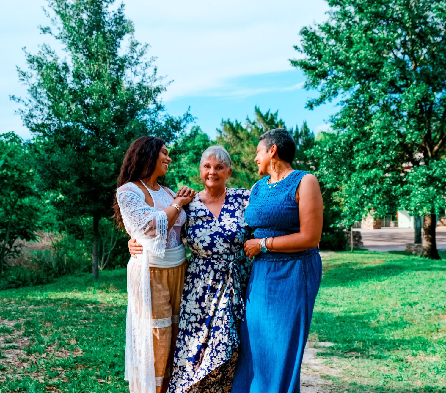 Three women laugh and embrace outdoors in a sunny green park during a joyful family photo session.