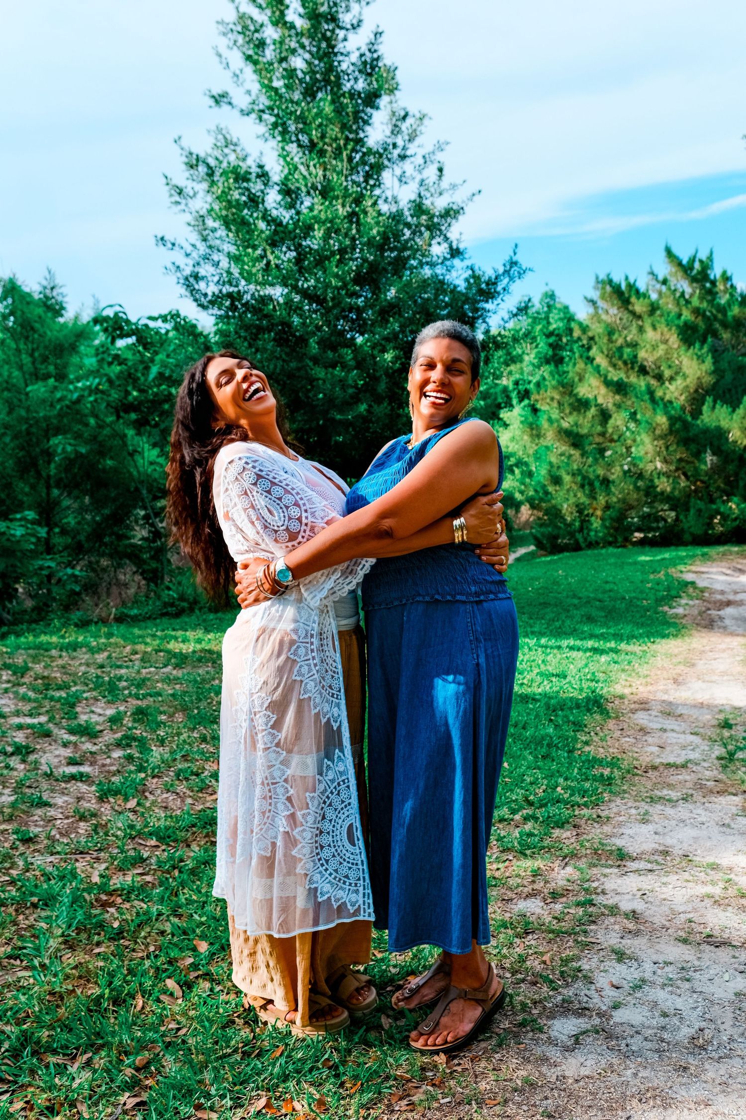 Two women share a warm embrace and laugh together on a sunny green lawn in casual summer outfits.
