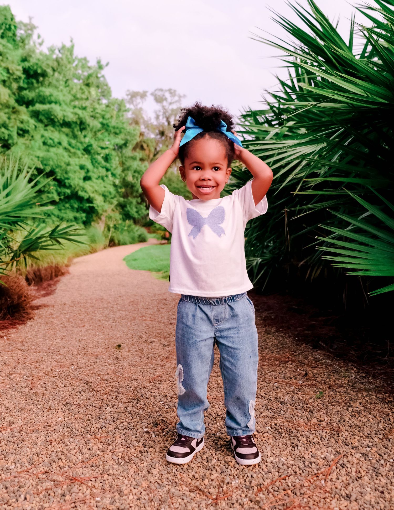 Toddler girl in blue bow, white tee, and jeans smiling on a garden path surrounded by lush greenery.