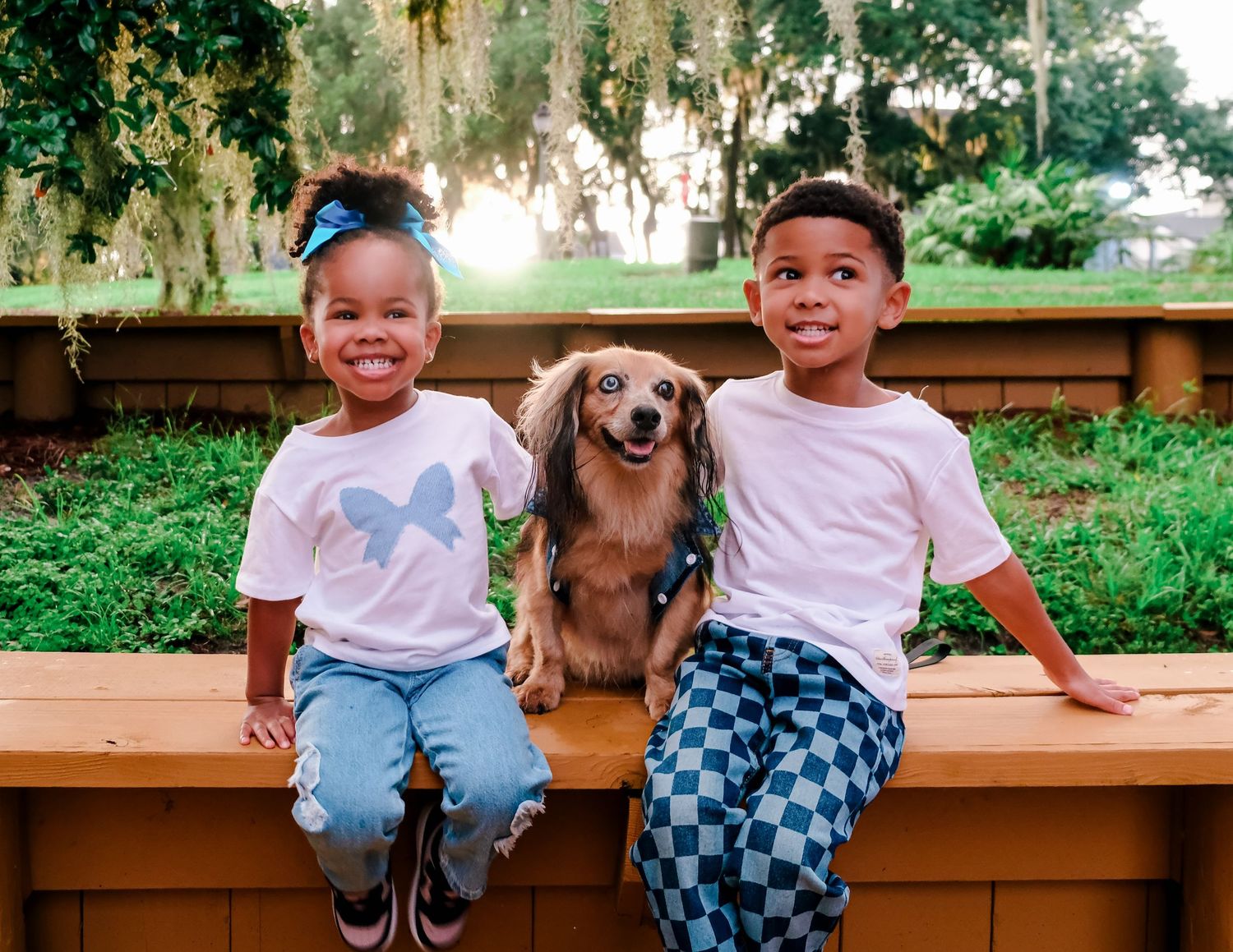 Two smiling children sit on a wooden bench with a dachshund dog between them, outdoors with green trees behind.