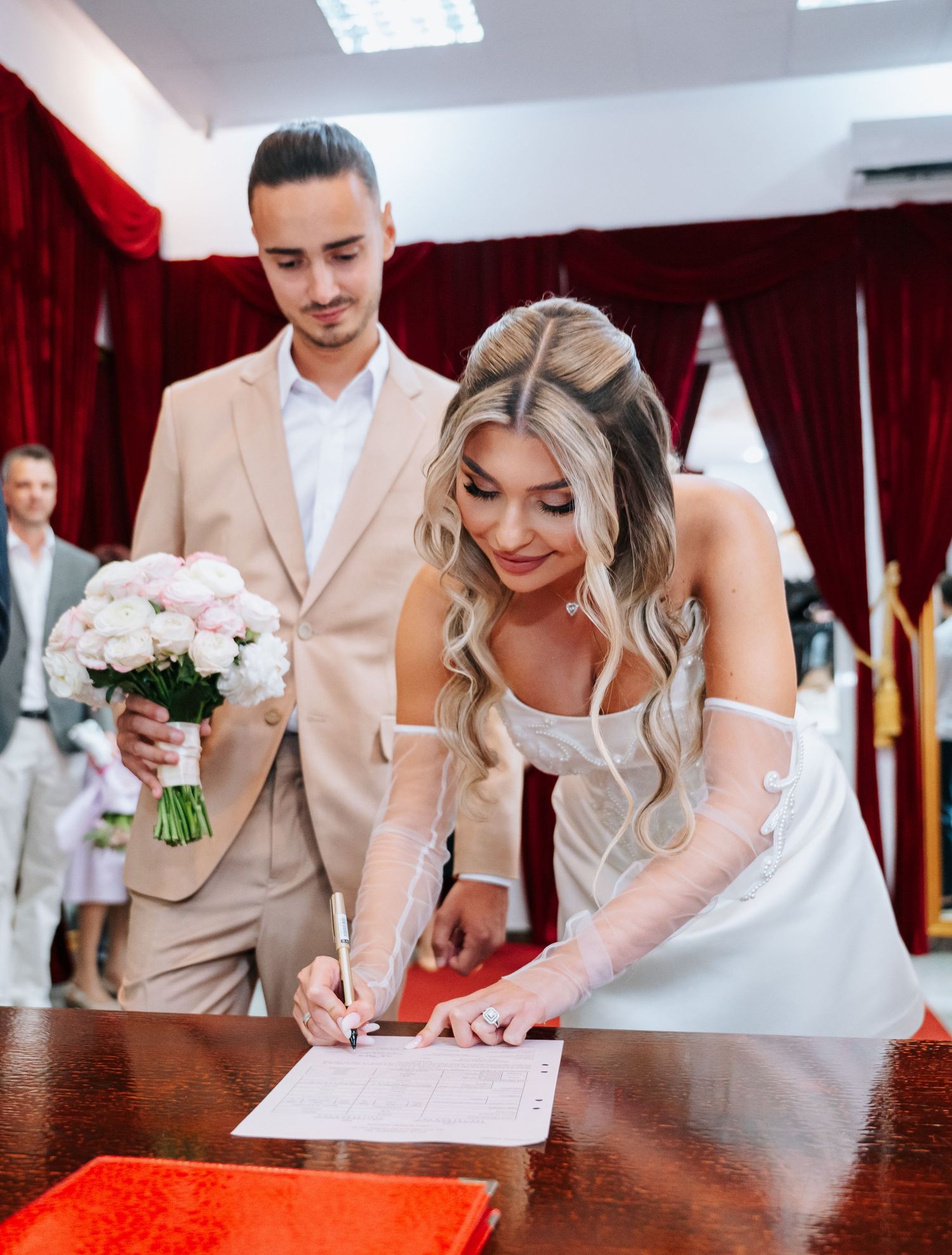 Bride signing wedding documents at ceremony while groom in beige suit holds pink flower bouquet.