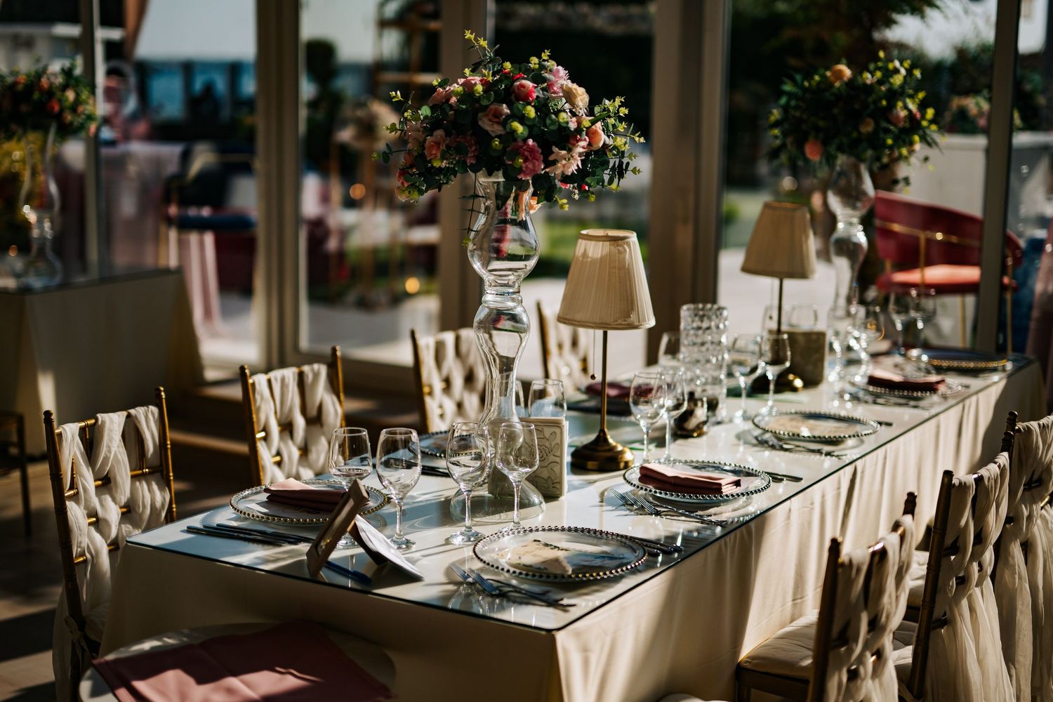 Elegant wedding reception table set with fine china, crystal glasses, floral centerpieces, and tufted chairs in a venue.