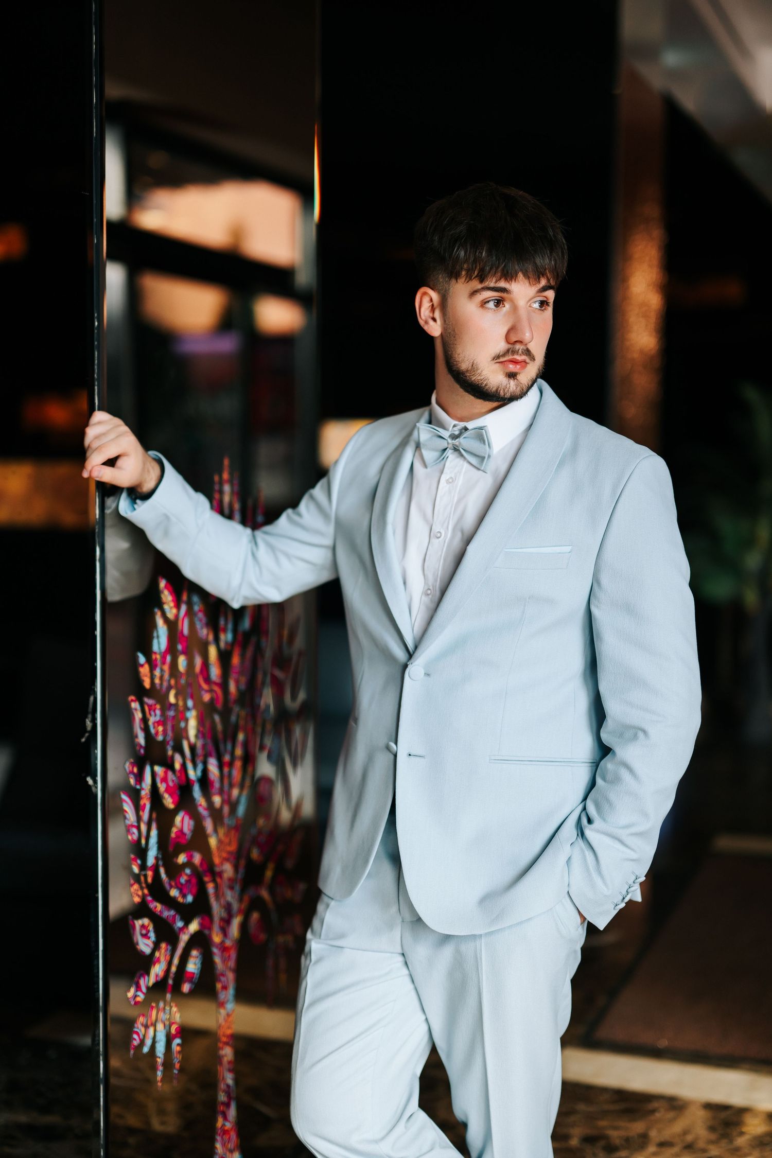 Young man in light blue suit and bow tie leaning against a dark door frame, looking to the side.