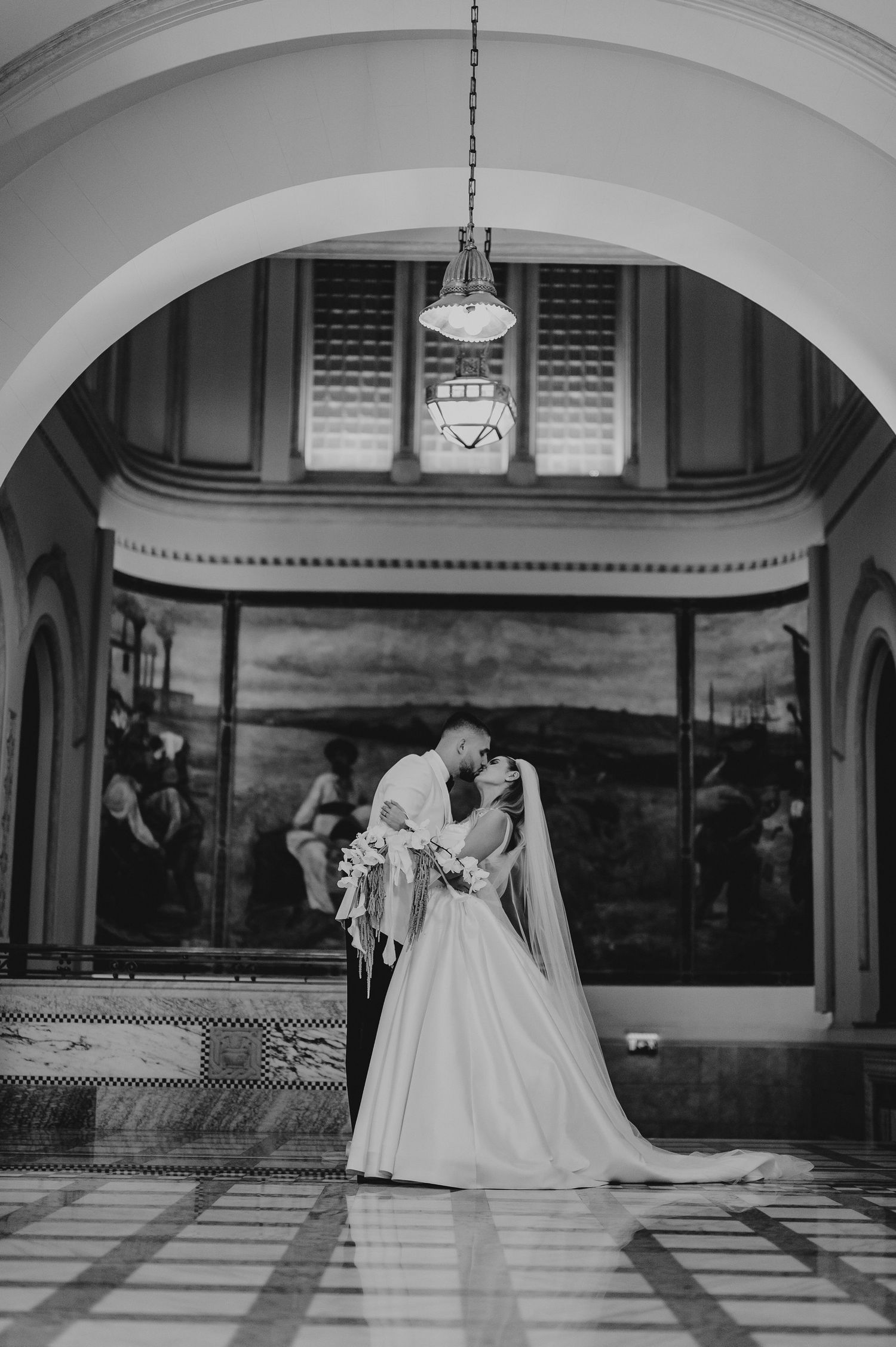 Black and white photo of newlyweds sharing a kiss under an ornate arch in a grand historic building.