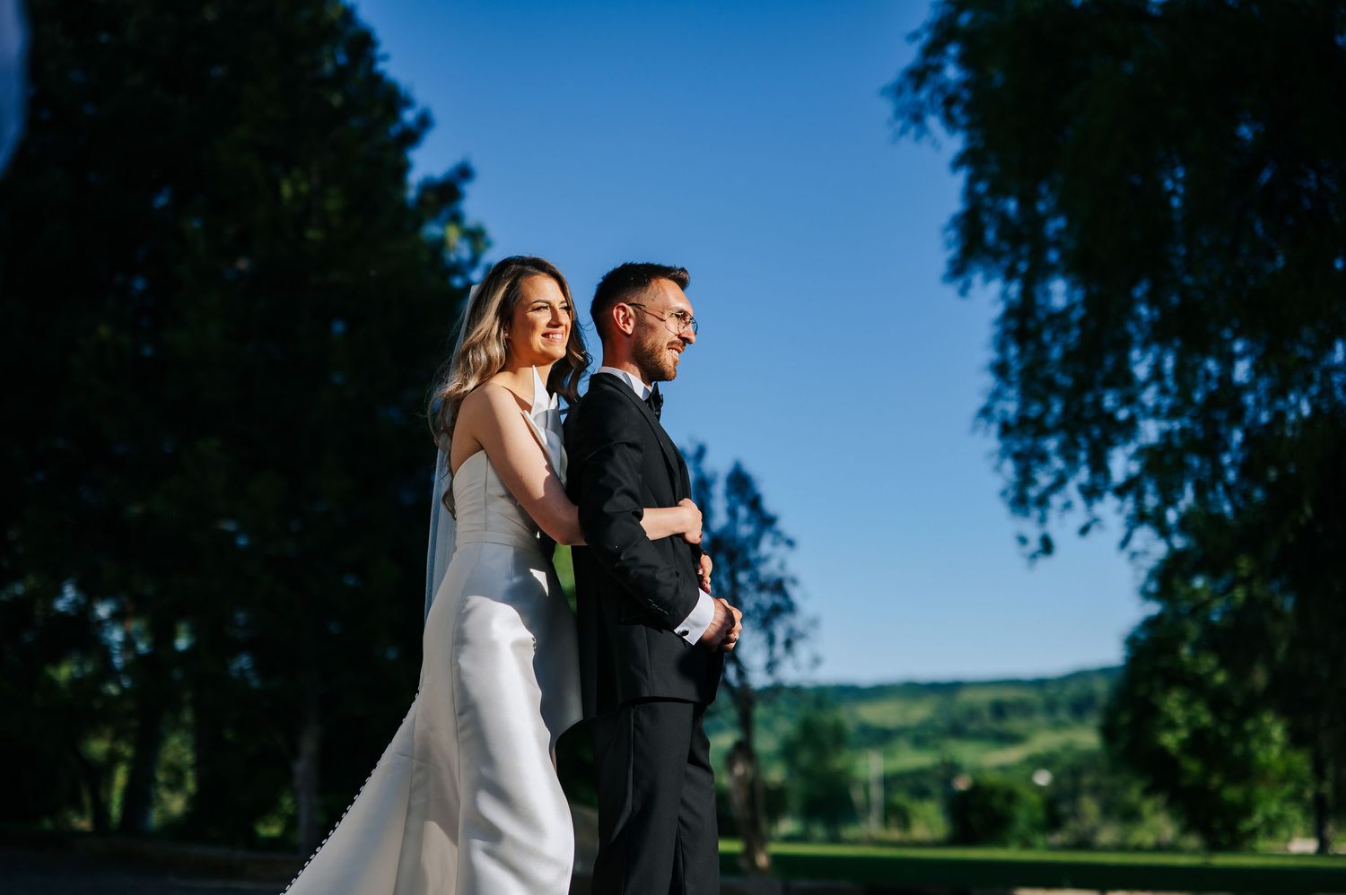 A bride and groom stand back-to-back outdoors on a sunny day, surrounded by lush green trees and rolling hills.