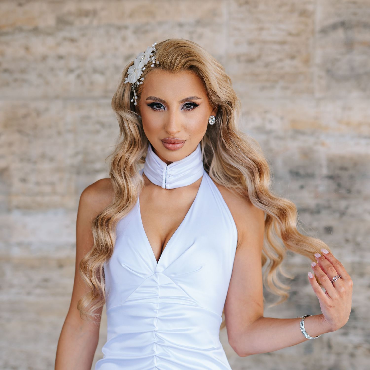 Woman in white halter dress with floral hair accessory and long wavy blonde hair poses against stone wall.