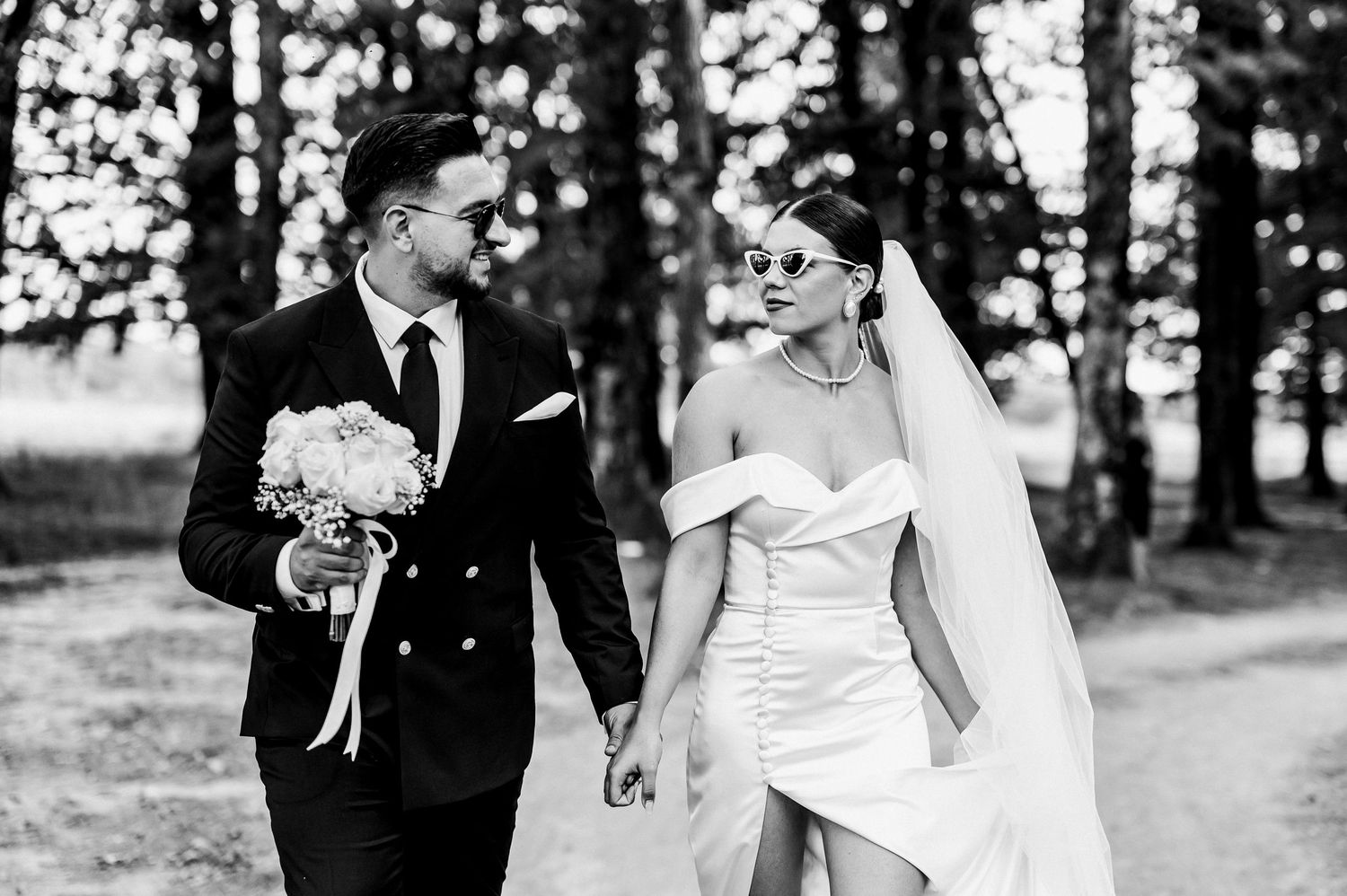Black and white photo of a bride and groom walking hand in hand outdoors, bride in strapless gown with veil.