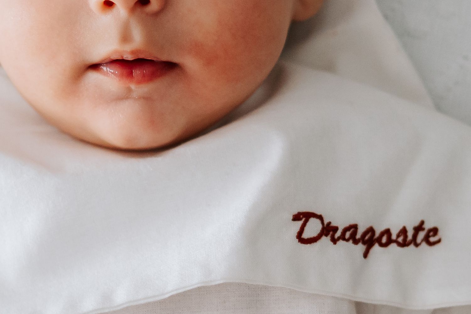 Close-up of newborn baby face resting on white fabric embroidered with the word Dragoste.