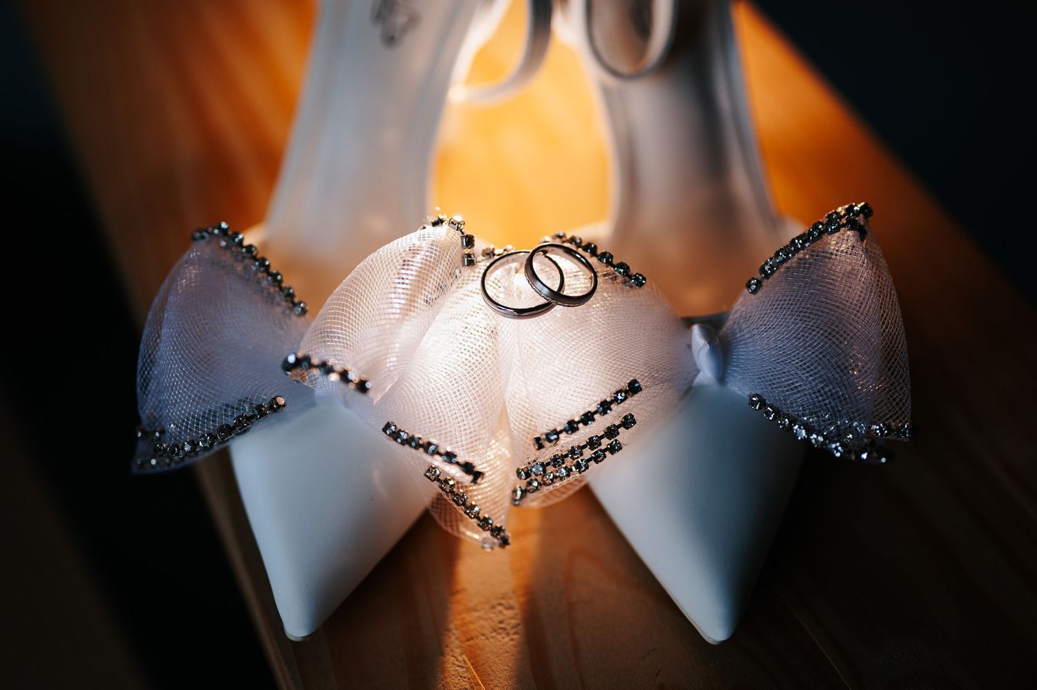 White bridal heels with black rhinestone details displaying two wedding rings nestled between the shoes.