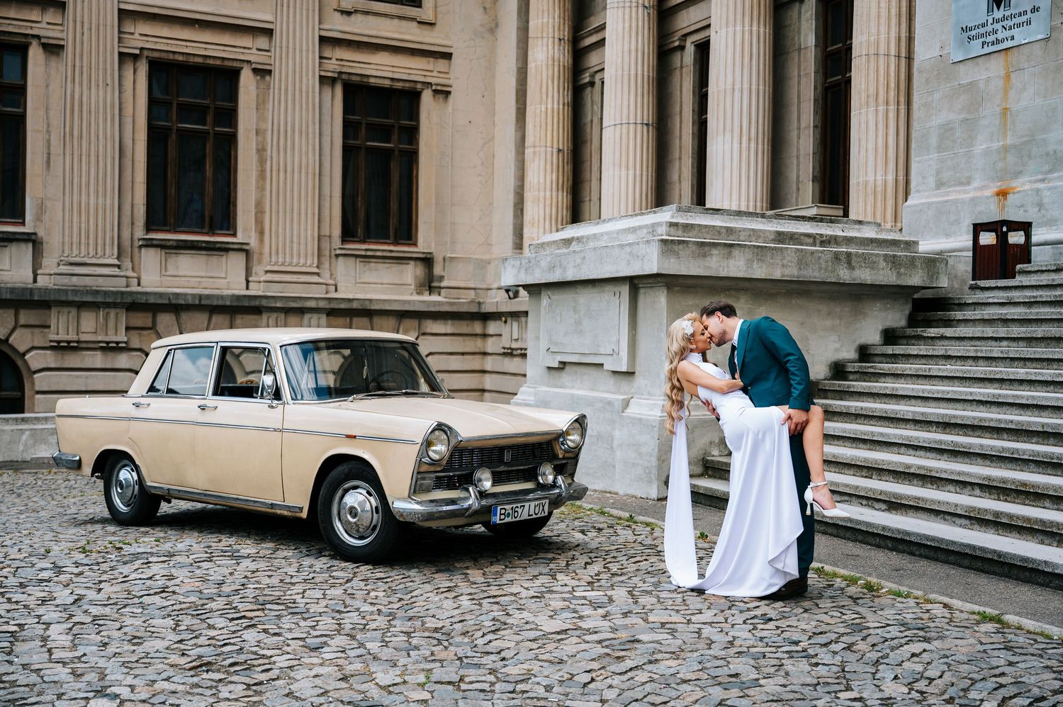 Bride and groom share a romantic dip kiss beside a vintage beige car on cobblestones outside a grand building.