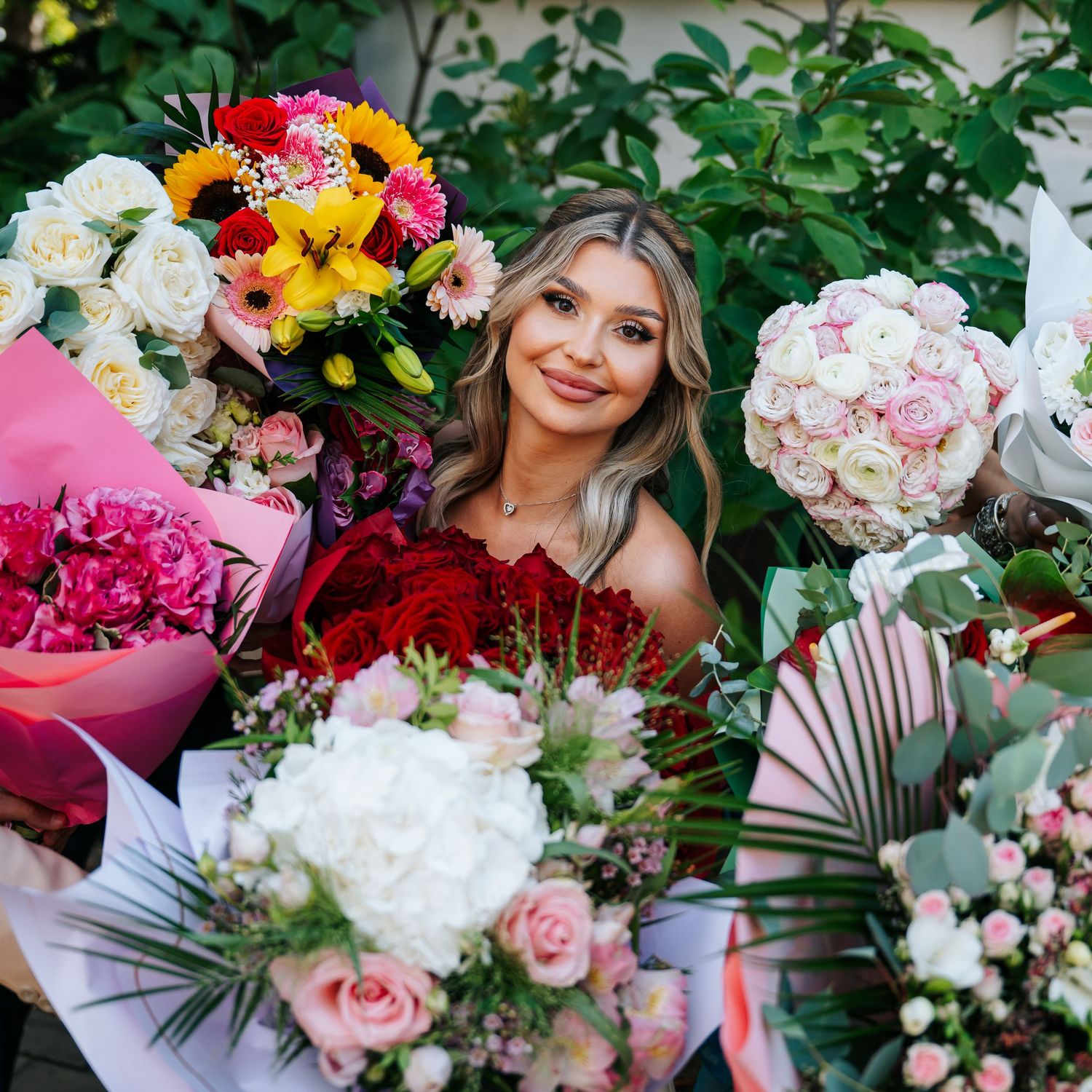 Smiling blonde woman surrounded by colorful flower bouquets including roses and gerberas outdoors.