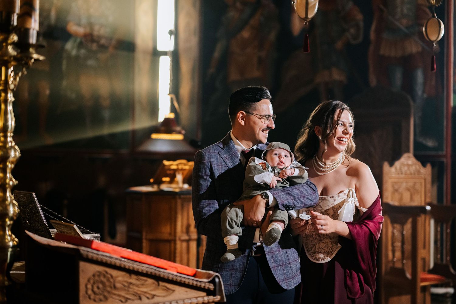 Smiling couple in elegant attire holds their baby inside a beautifully ornate Orthodox church interior.
