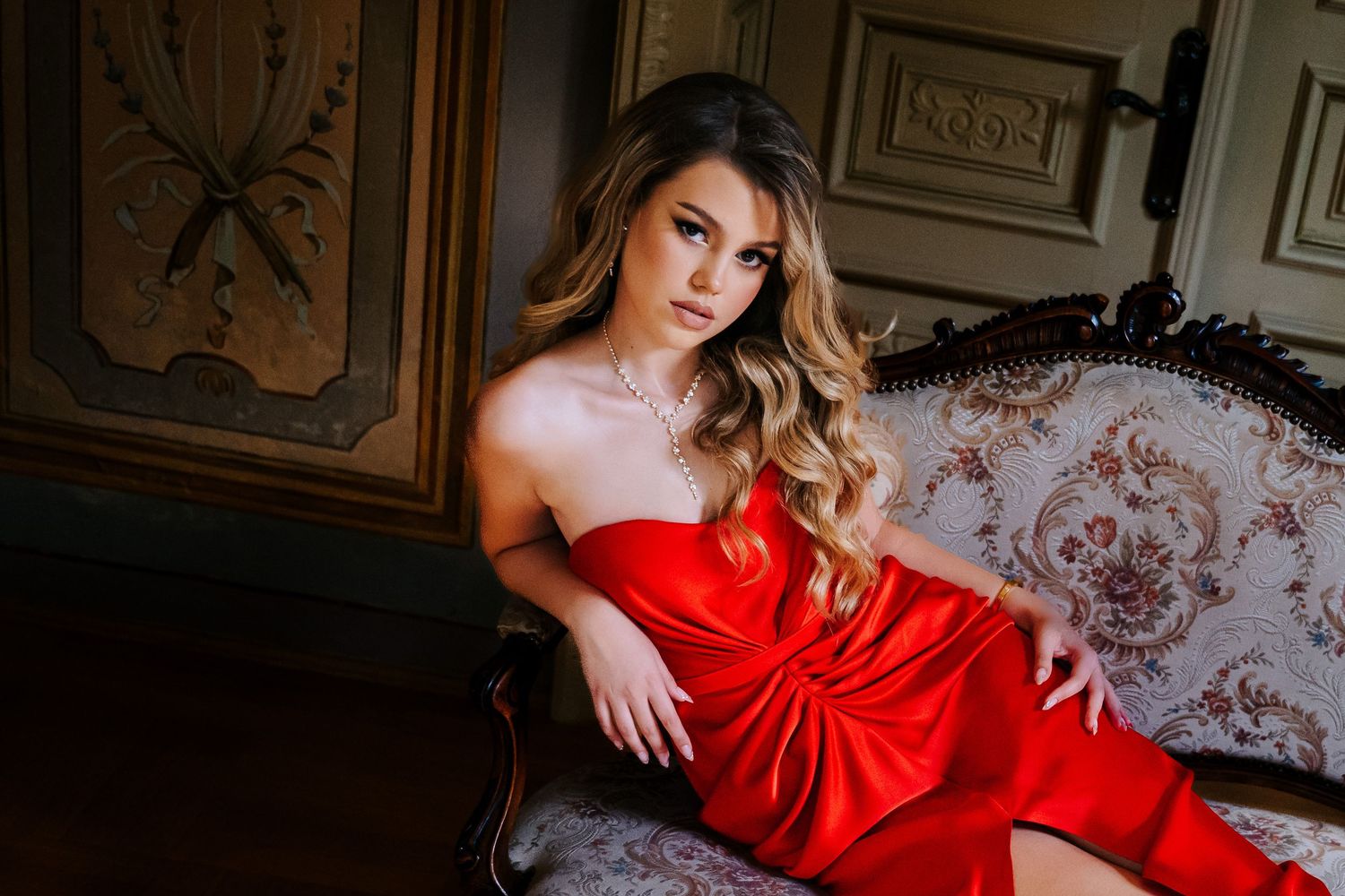 Young woman in elegant red strapless dress sitting gracefully on vintage ornate sofa in classic interior.
