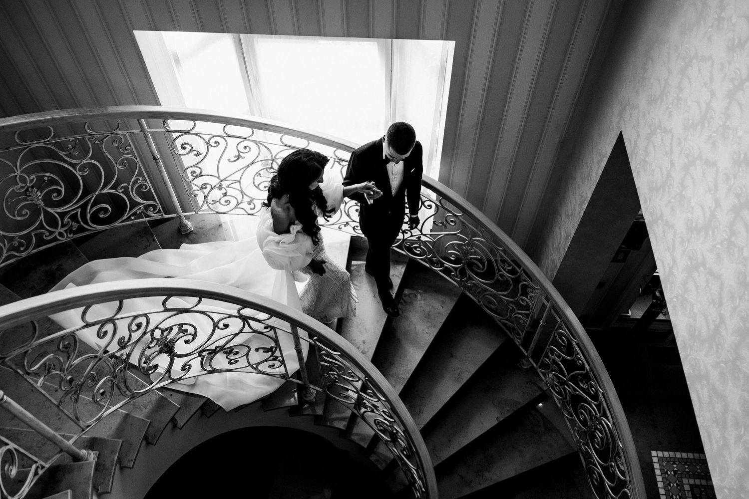 Aerial black and white photo of bride and groom descending elegant spiral staircase with ornate iron railing.