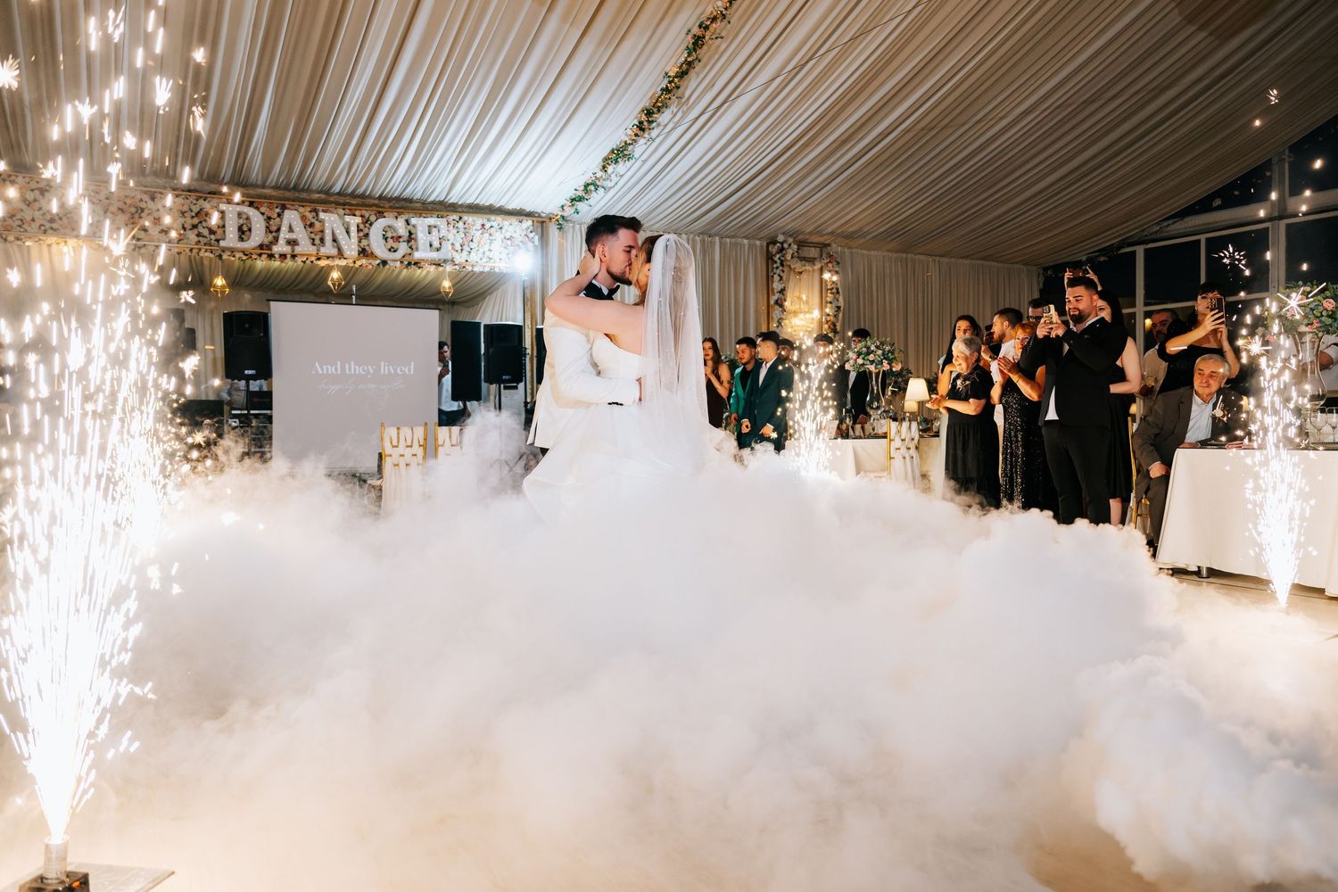 Bride and groom share first dance on a cloud of fog with sparkling fireworks and DANCE sign at elegant wedding reception.
