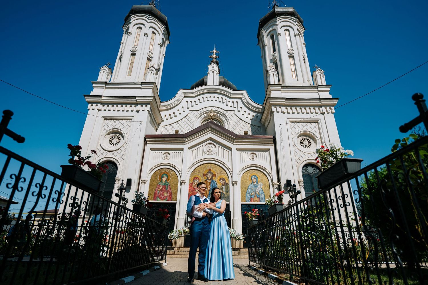 Couple in formal attire poses with baby before ornate white Orthodox church with twin towers under blue sky.