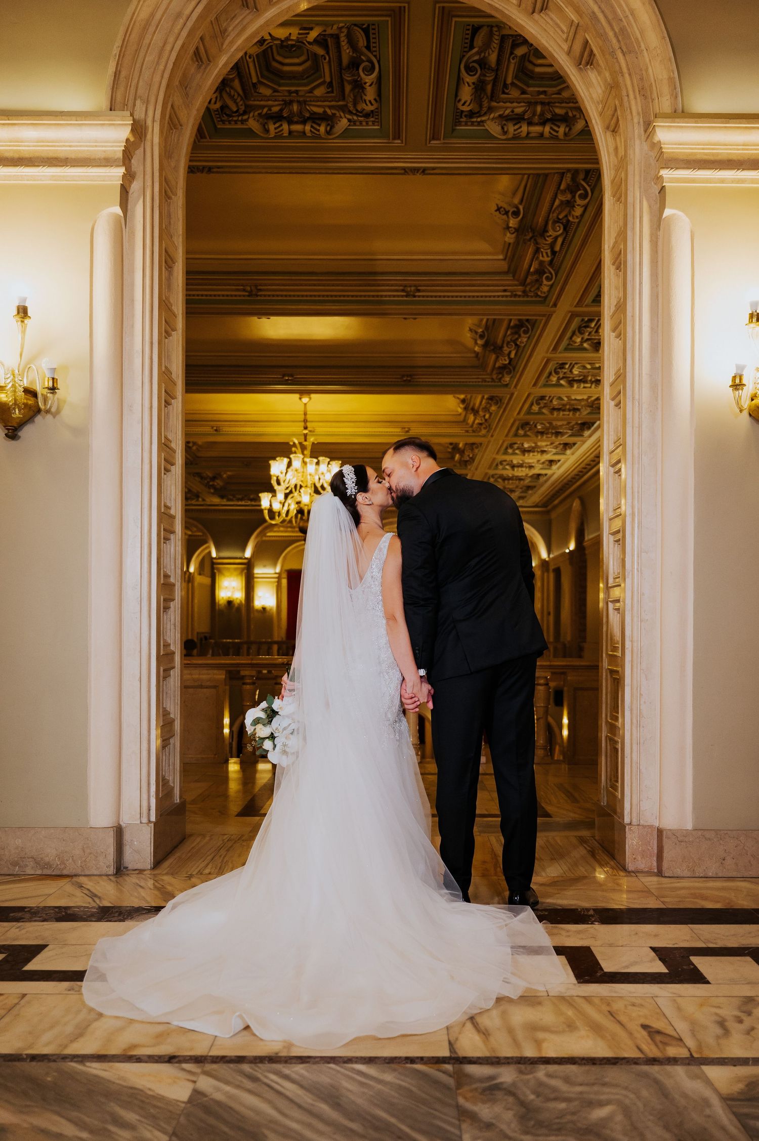 Bride and groom share a kiss in an ornate golden hall, her white gown and veil trailing elegantly on marble floors.