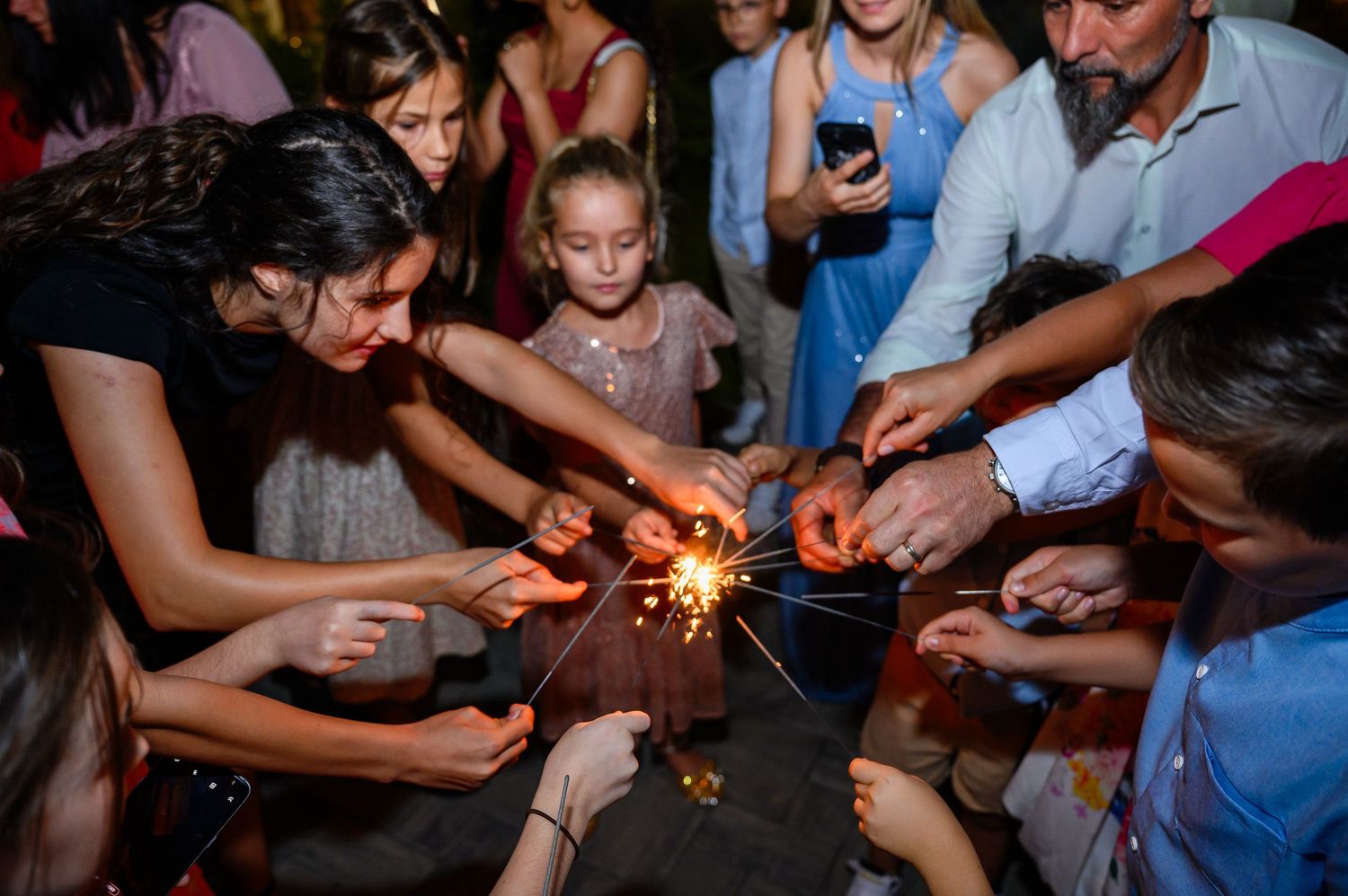 Adults and children gather around sparklers at a festive nighttime celebration party.