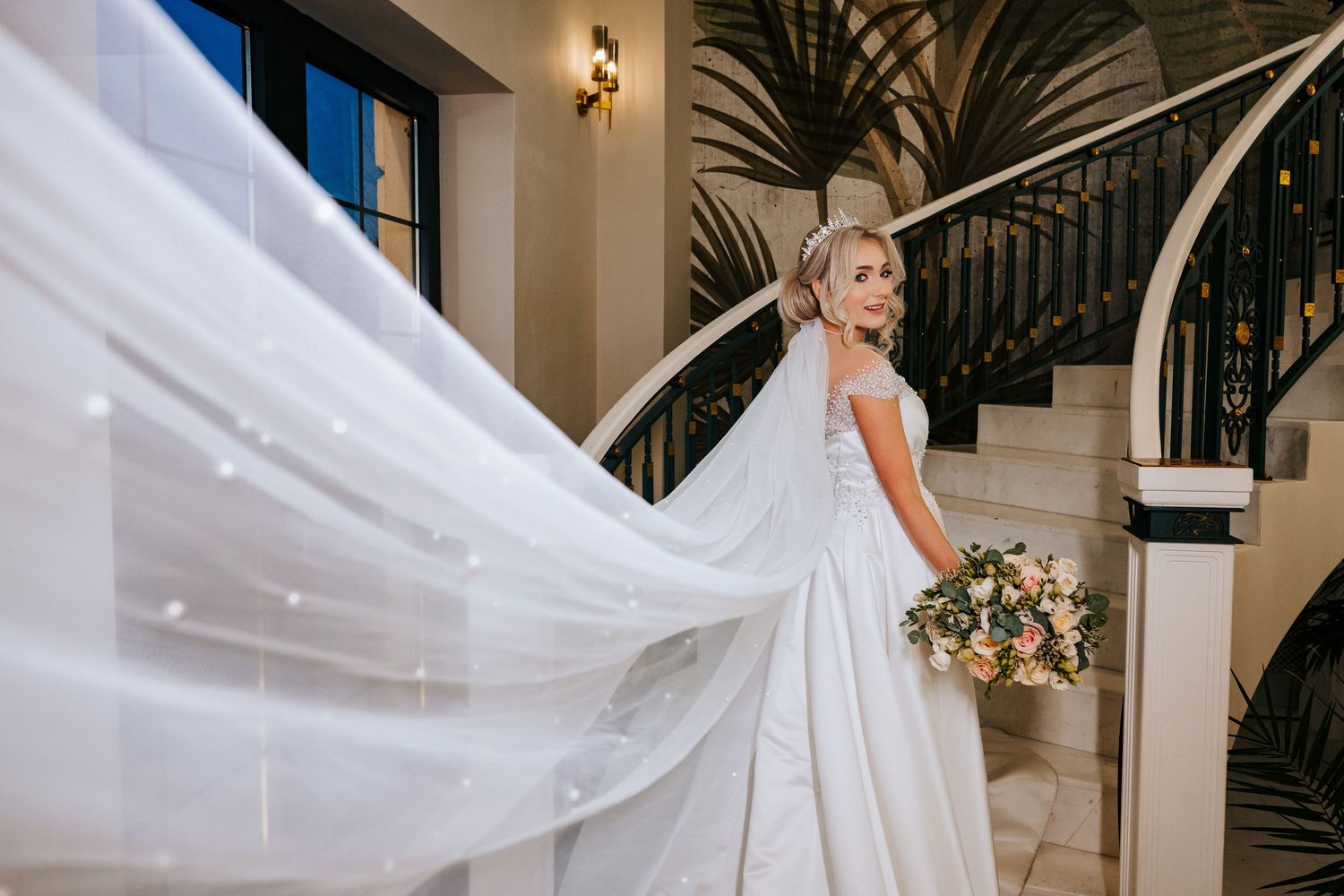 Bride in a flowing white gown with a billowing veil holds a bouquet near an elegant staircase.
