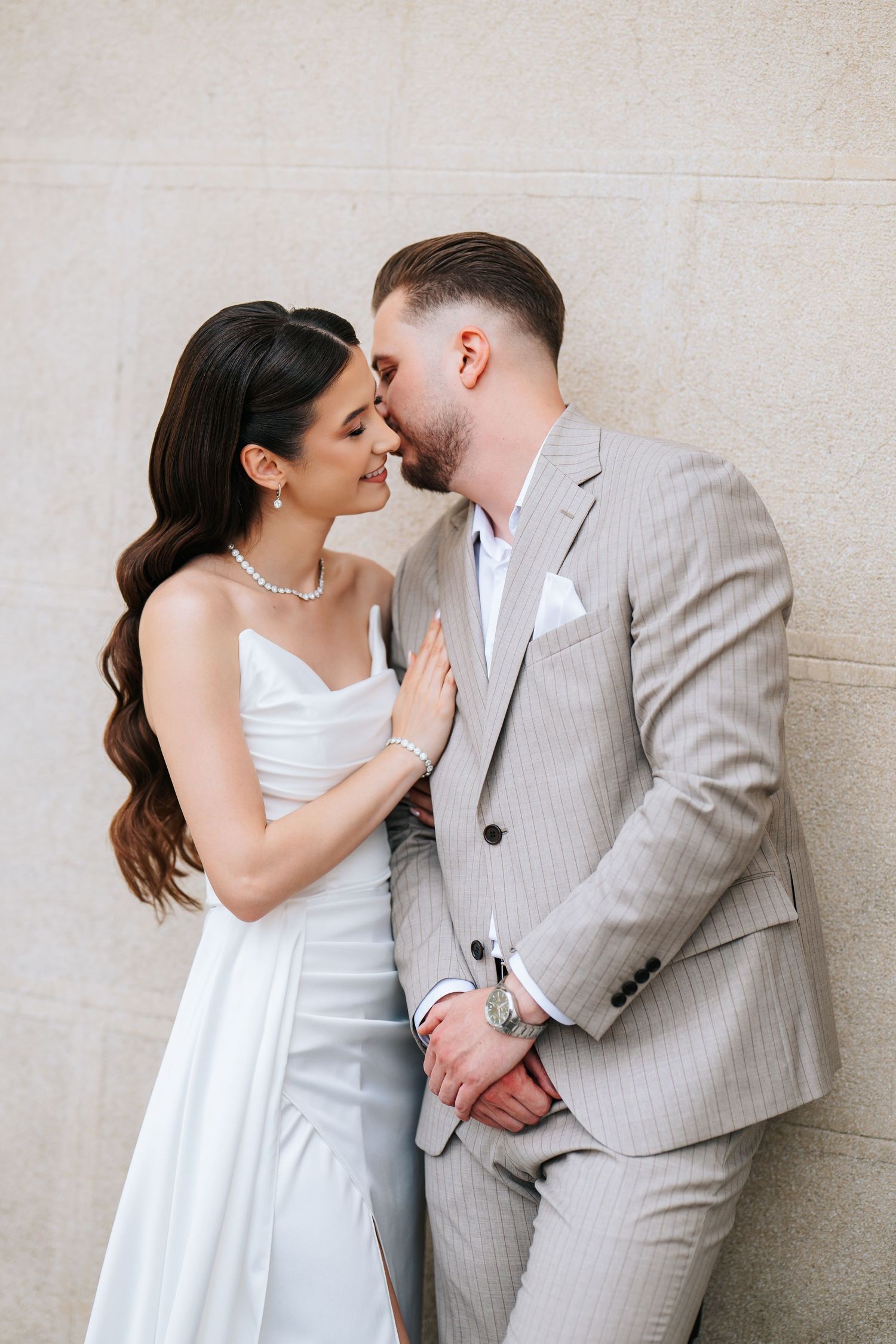 Bride in white gown and pearl necklace leans close to groom in striped suit against stone wall.
