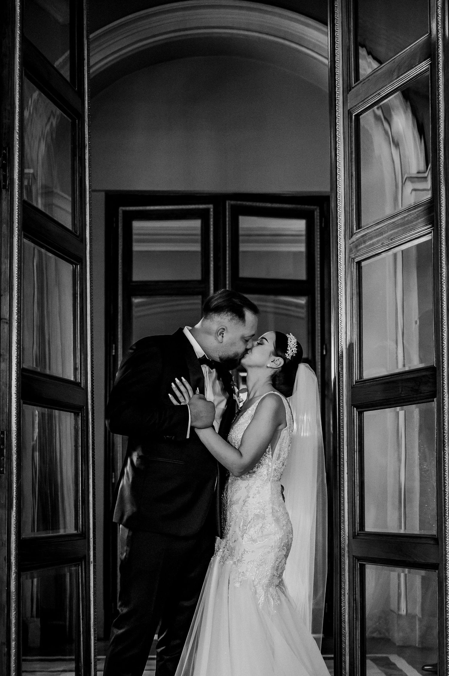 Black and white wedding photo of bride and groom sharing a kiss in an elegant doorway, bride in lace gown with veil.