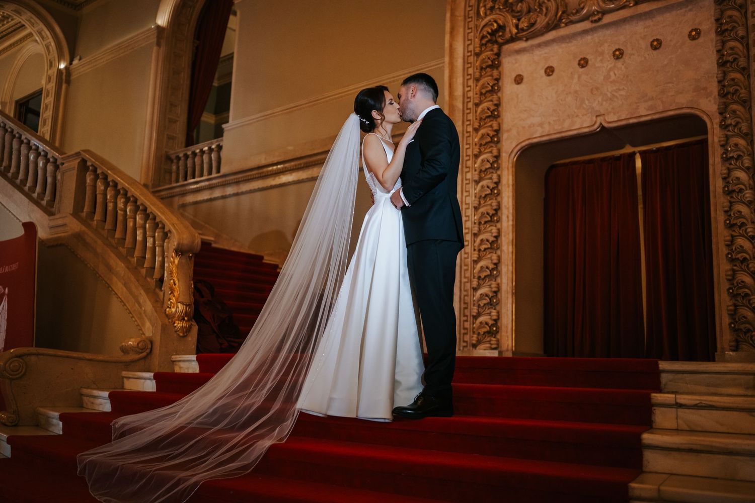 Bride and groom share a romantic kiss on grand red-carpeted staircase in elegant wedding venue.