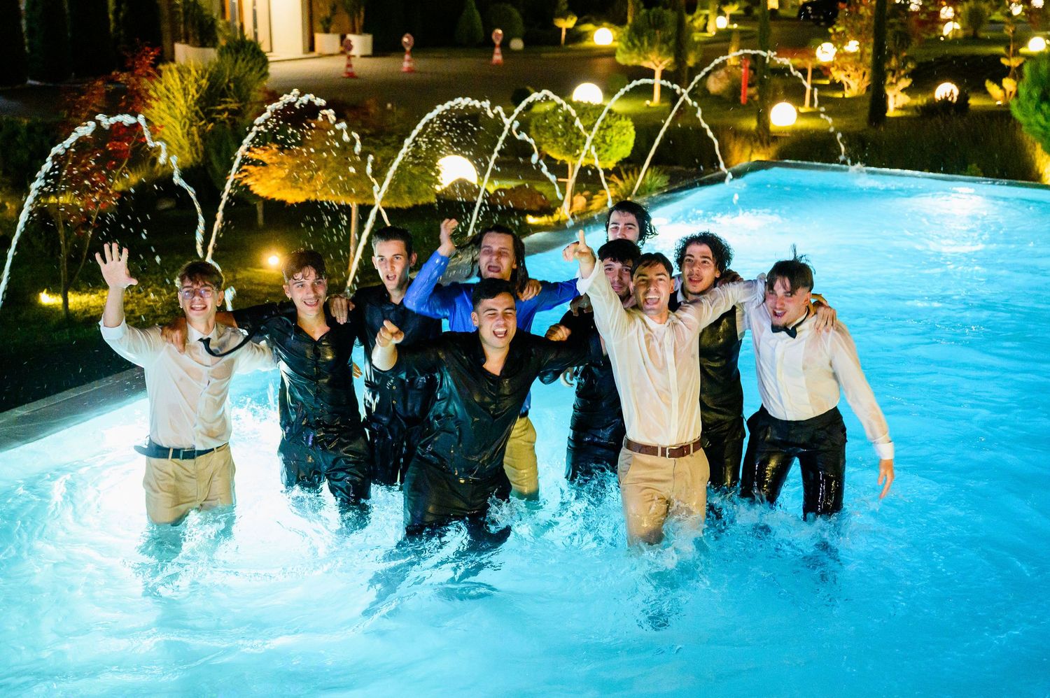 Group of friends celebrating in illuminated pool at night with water fountains splashing around them.