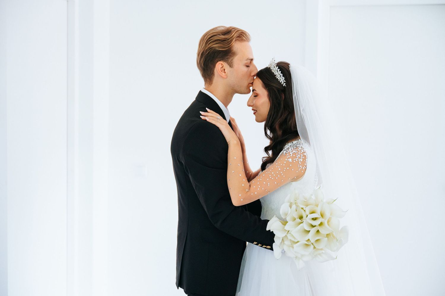 Bride and groom in wedding attire share a romantic embrace, she holds white calla lily bouquet against white backdrop.