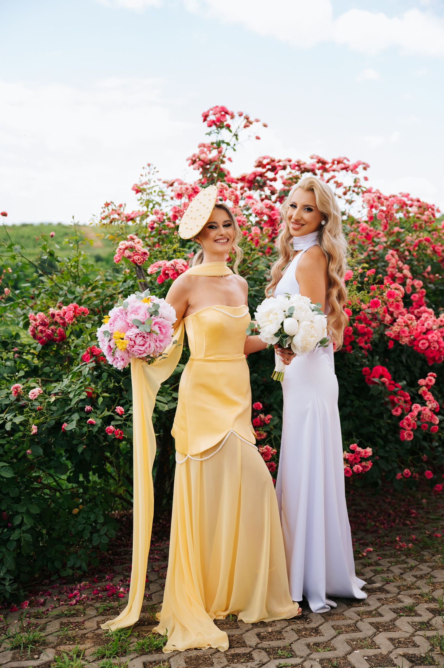 Two elegant women in yellow and white gowns holding floral bouquets pose before blooming pink rose bushes outdoors.