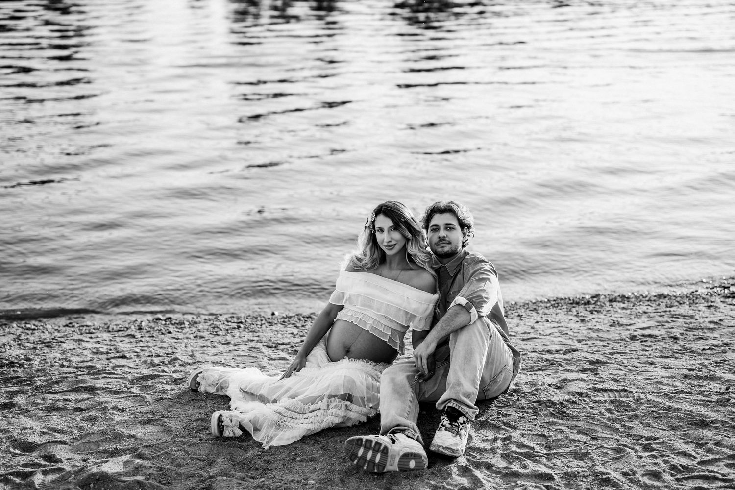 Black and white photo of a couple sitting on a rocky shoreline, woman in flowy dress showing baby bump.