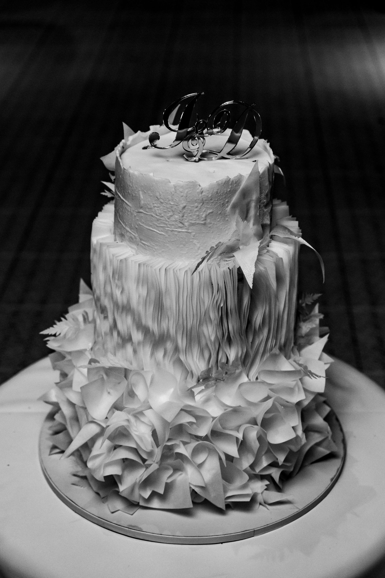 Black and white photo of elegant three-tier wedding cake with ruffled fondant base and decorative topper.