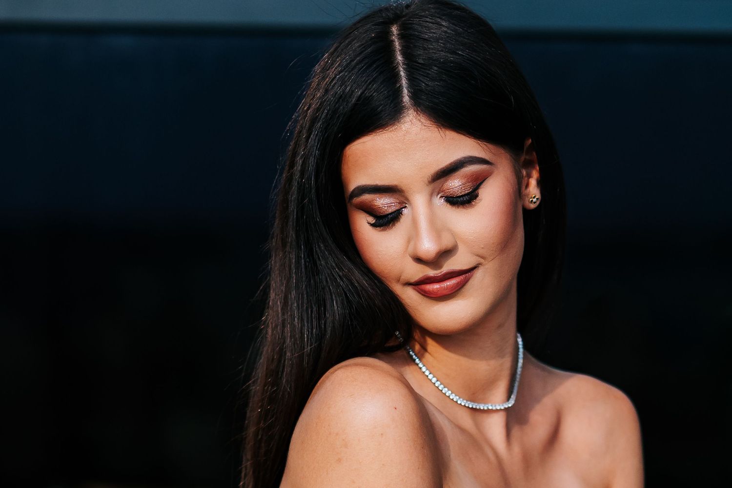 Young woman with long dark hair, pearl necklace, and elegant makeup poses gracefully with a soft downward gaze.
