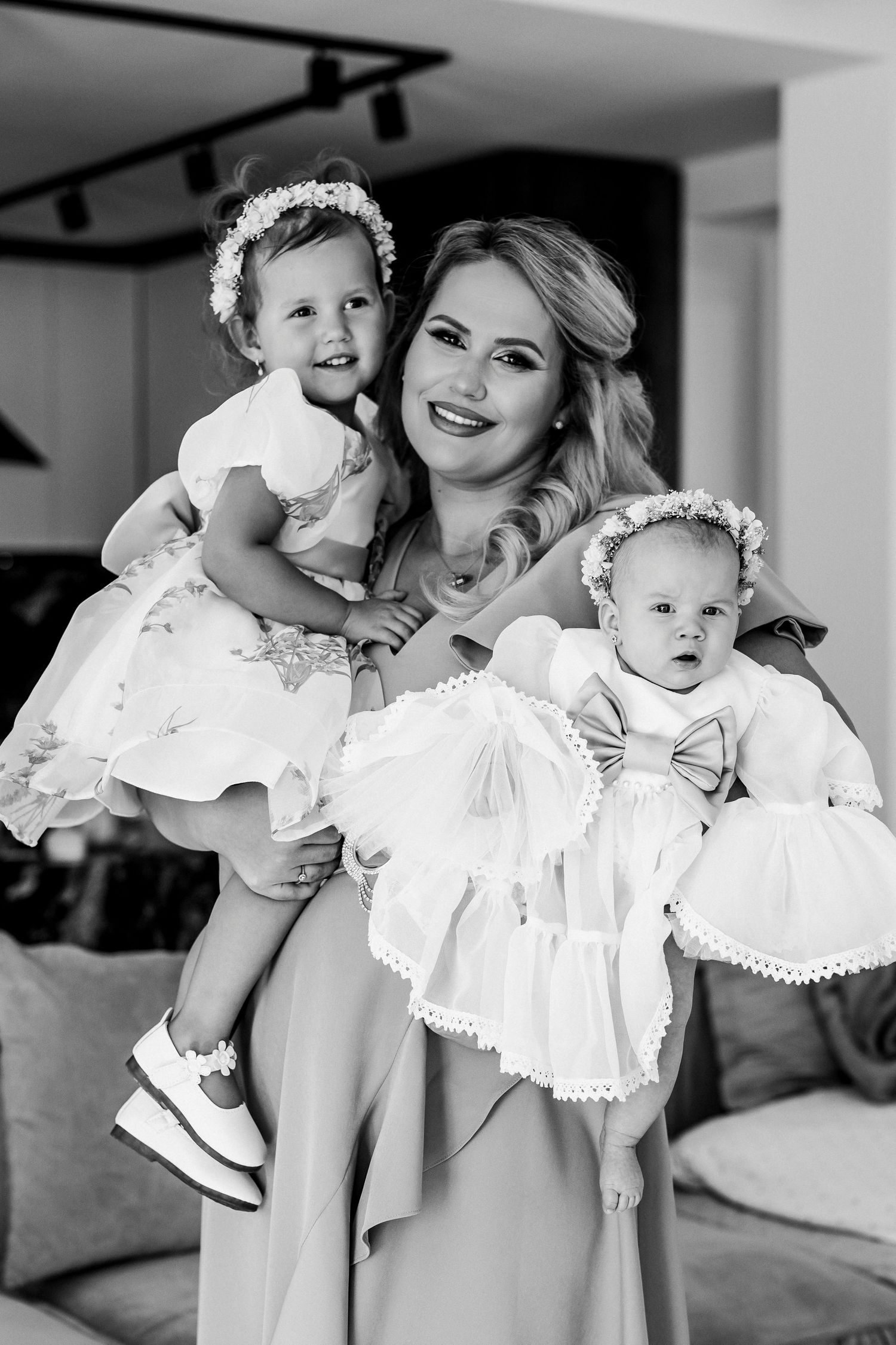 Black and white photo of a smiling woman holding two young girls in white dresses and floral headbands indoors.