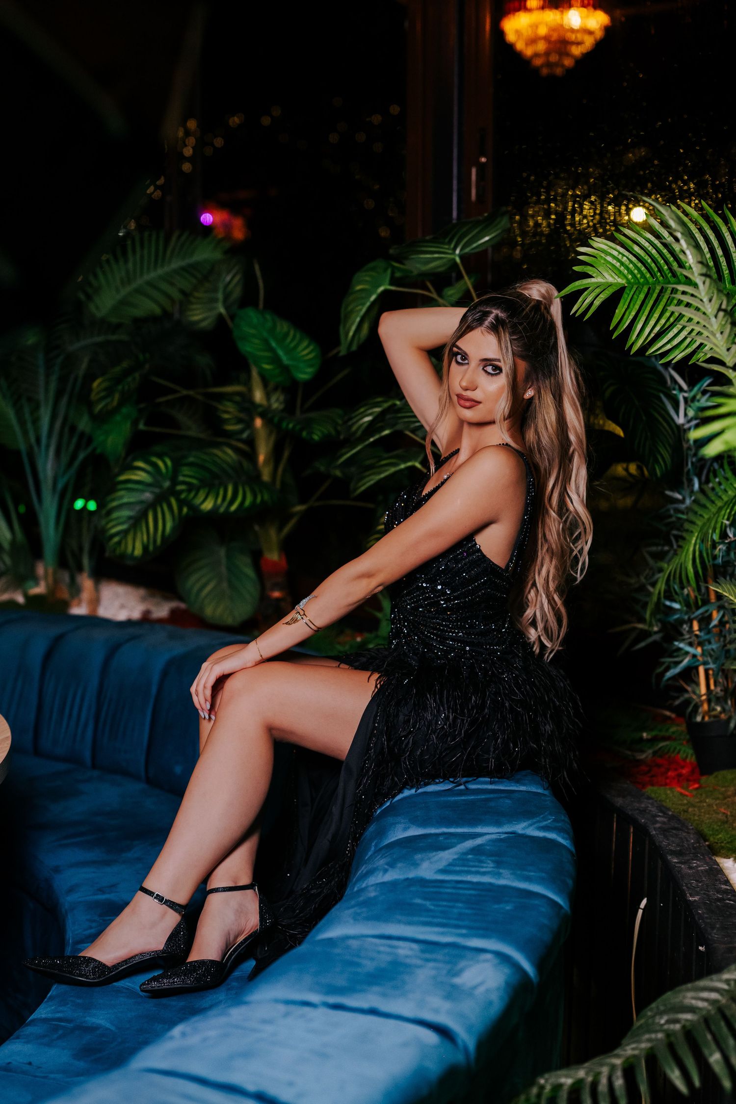 Woman in black sequin feather dress seated on blue velvet sofa amid tropical plants.