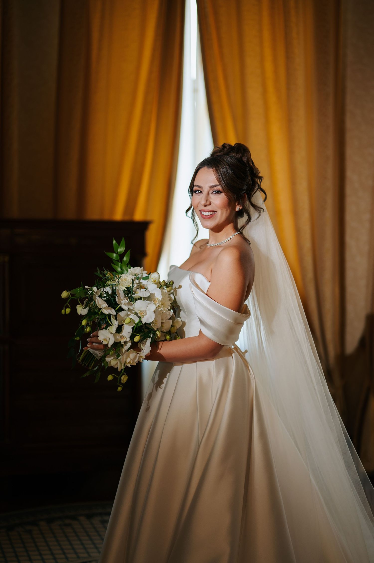 Elegant bride in satin off-shoulder gown holding white bouquet beside golden curtains.