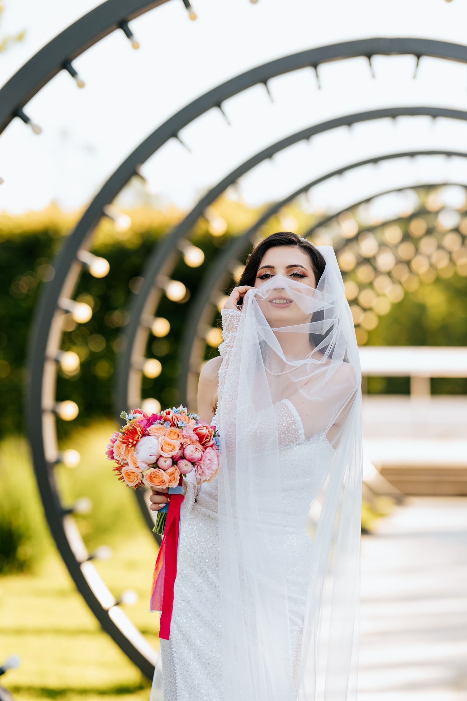 Elegant bride in white gown holding colorful bouquet with red ribbon, posing under circular arch with lights outdoors.