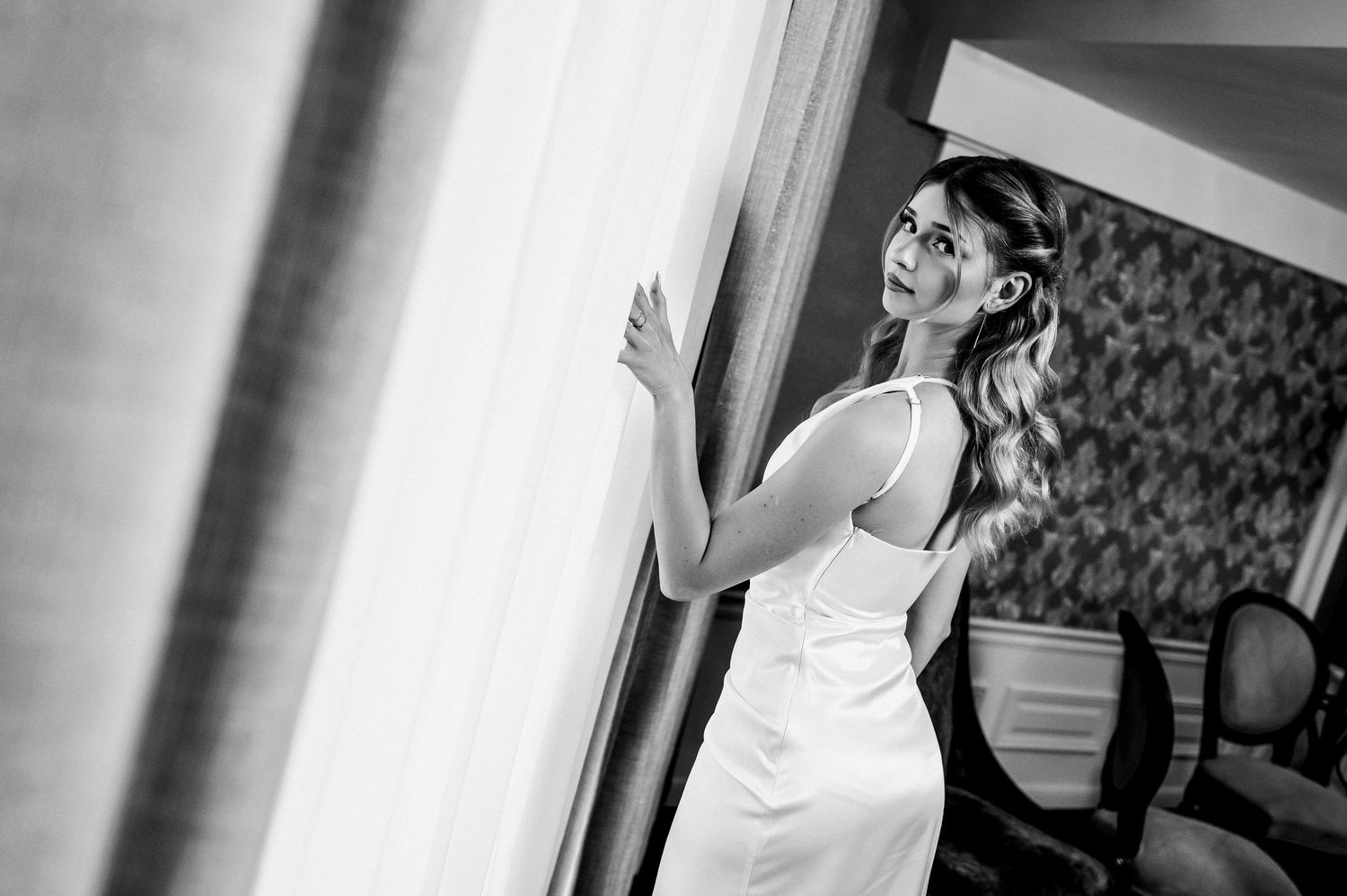 Bride in white dress standing by window in black and white wedding photo.