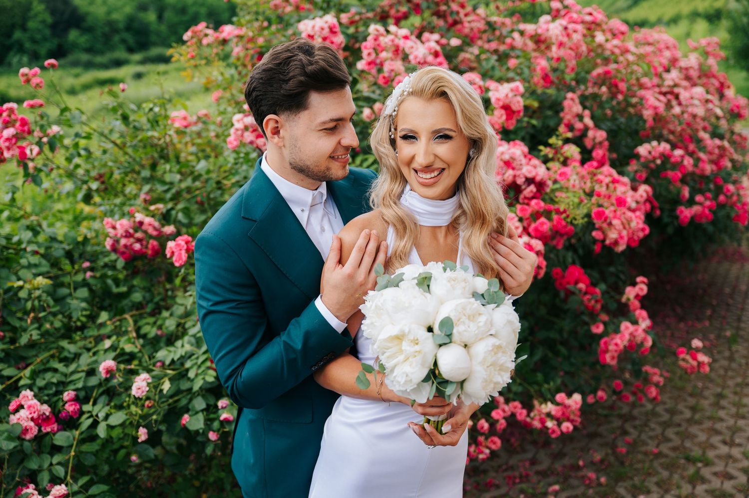 Bride and groom embracing in front of pink roses, bride holding white peony bouquet in elegant wedding attire.